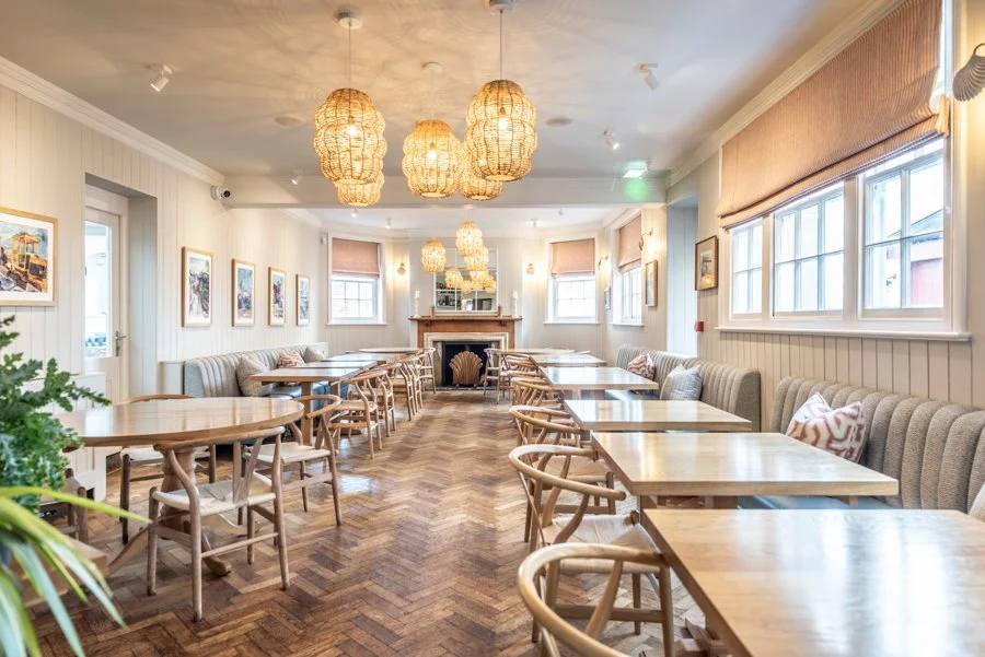 A cozy dining room with wooden tables and chairs, beige cushioned seating along the wall, a fireplace at the far end, large windows with curtains, and wicker pendant lights hanging from the ceiling.
