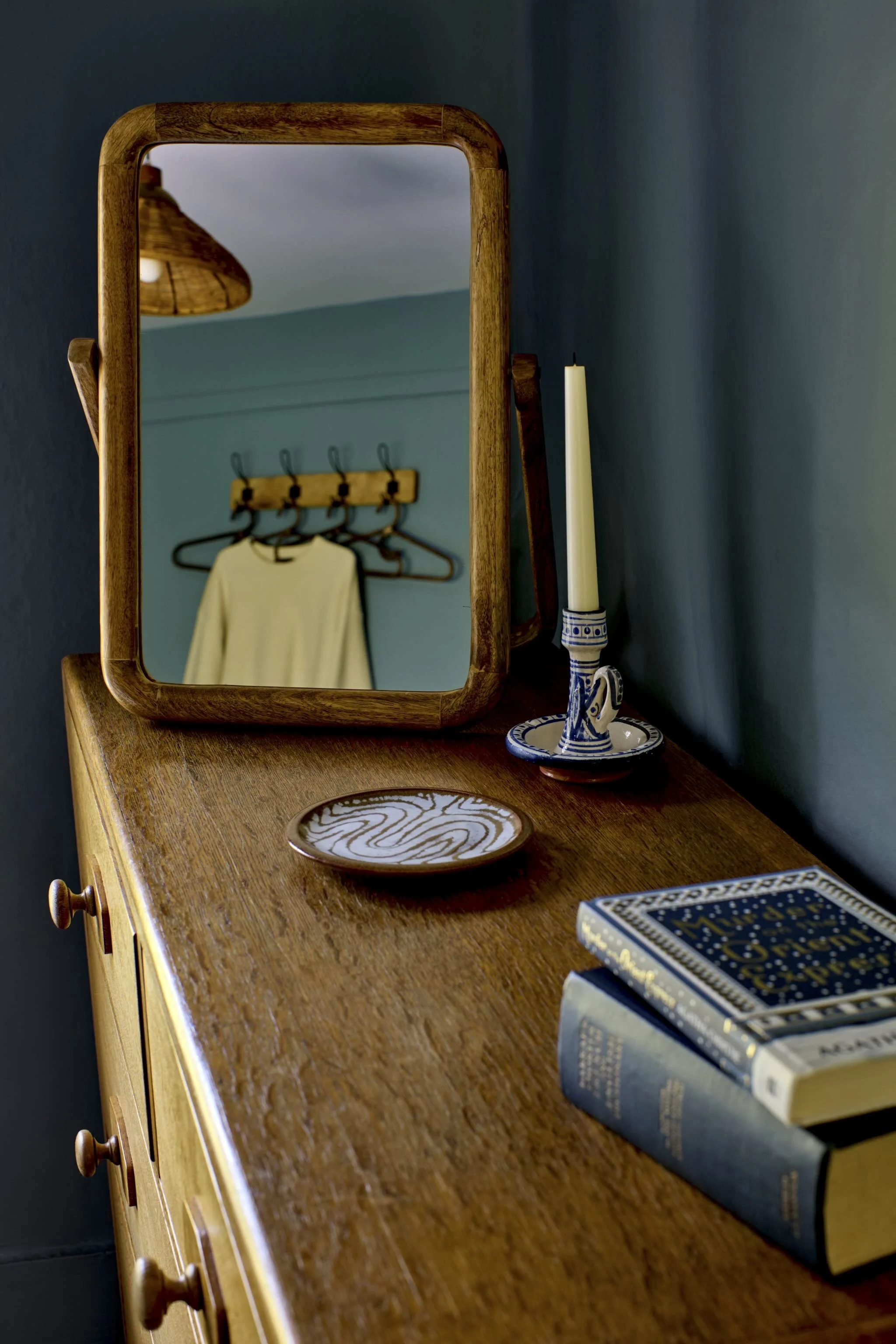 A wooden dresser with a mirror, a ceramic candle holder with a tall candle, a decorative plate, and a closed book on top.
