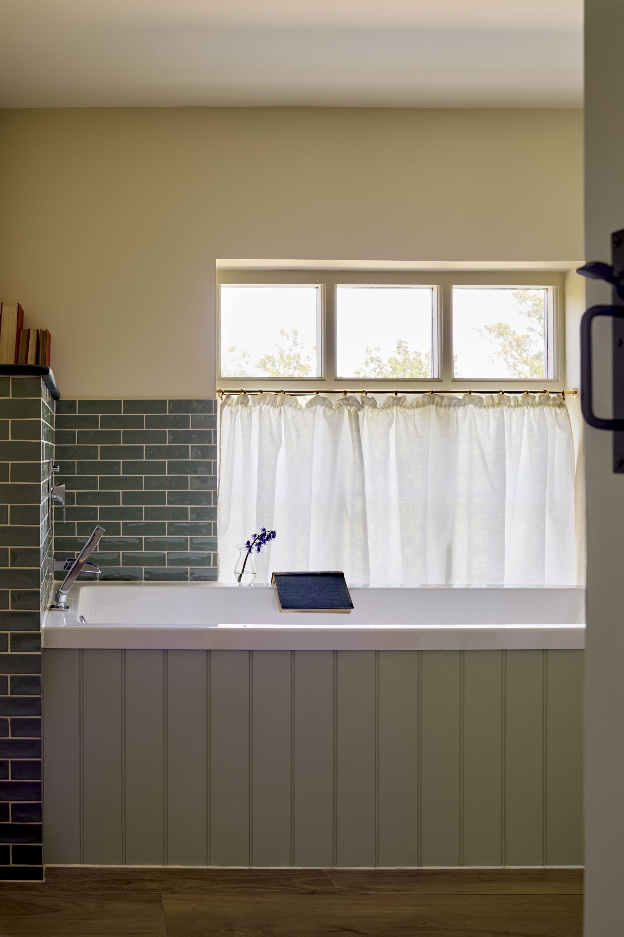 A cozy bathroom with a bathtub, green subway tile on the wall, and a large window with white curtains allowing natural light to fill the space.