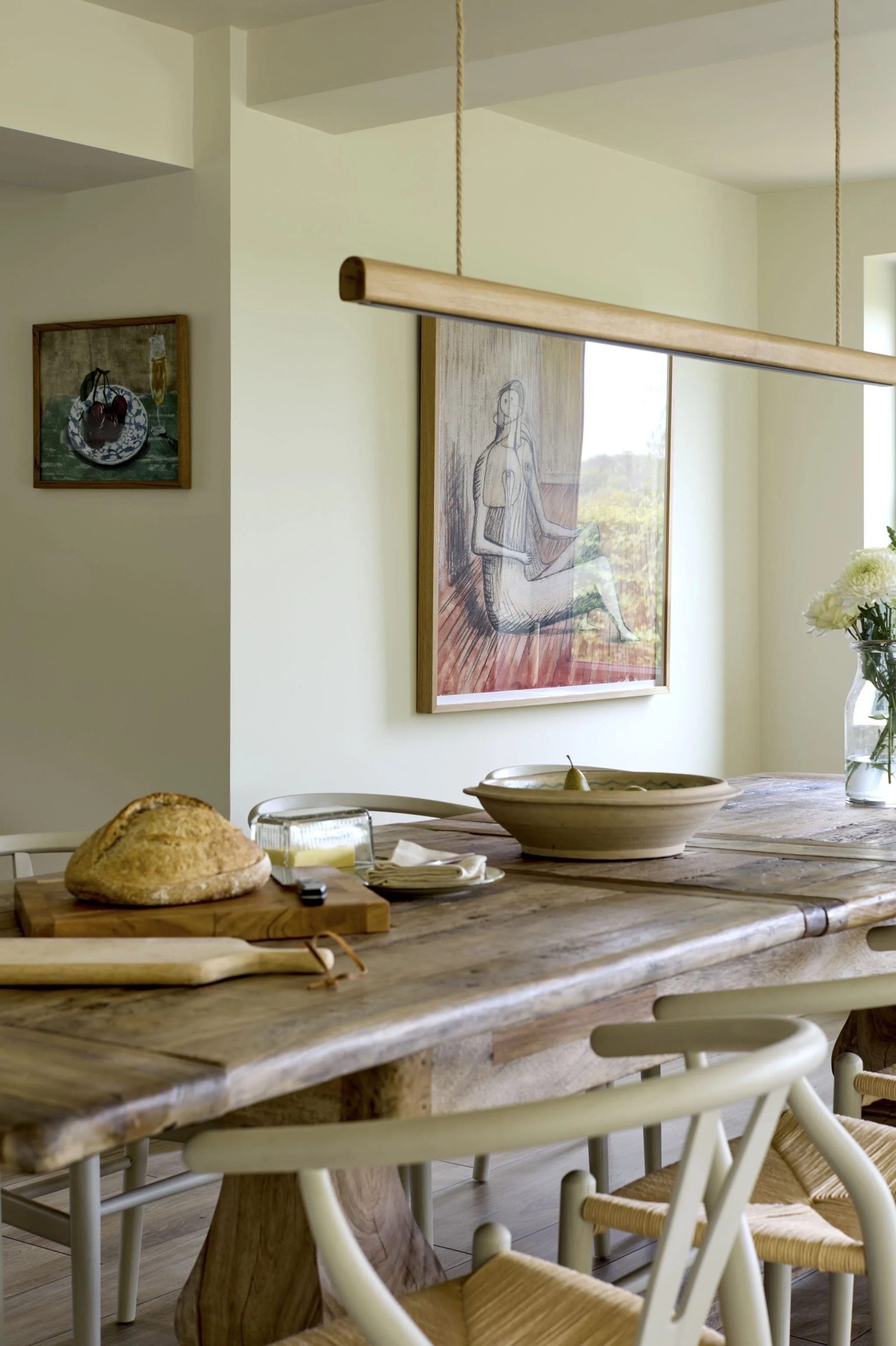 A dining area with a rustic wooden table, white chairs with woven seats, a loaf of bread on a cutting board, a bowl with a pear, and a windowed wall with artwork, including a framed artistic sketch of a seated woman.