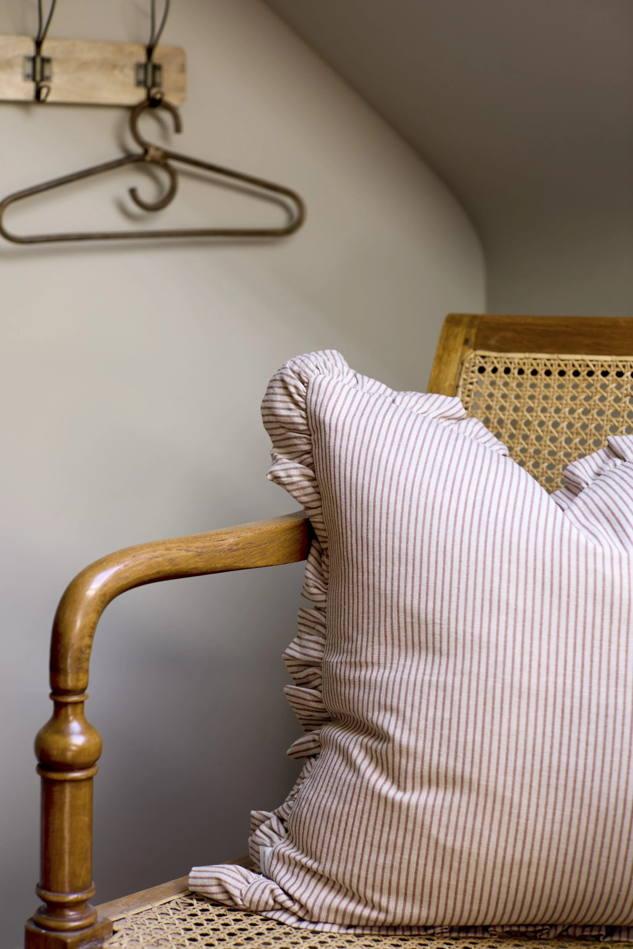 A wooden chair with a woven cane seat, a pink and white striped pillow with ruffled edges on it, and a beige wall with metal hangers and a wooden bar