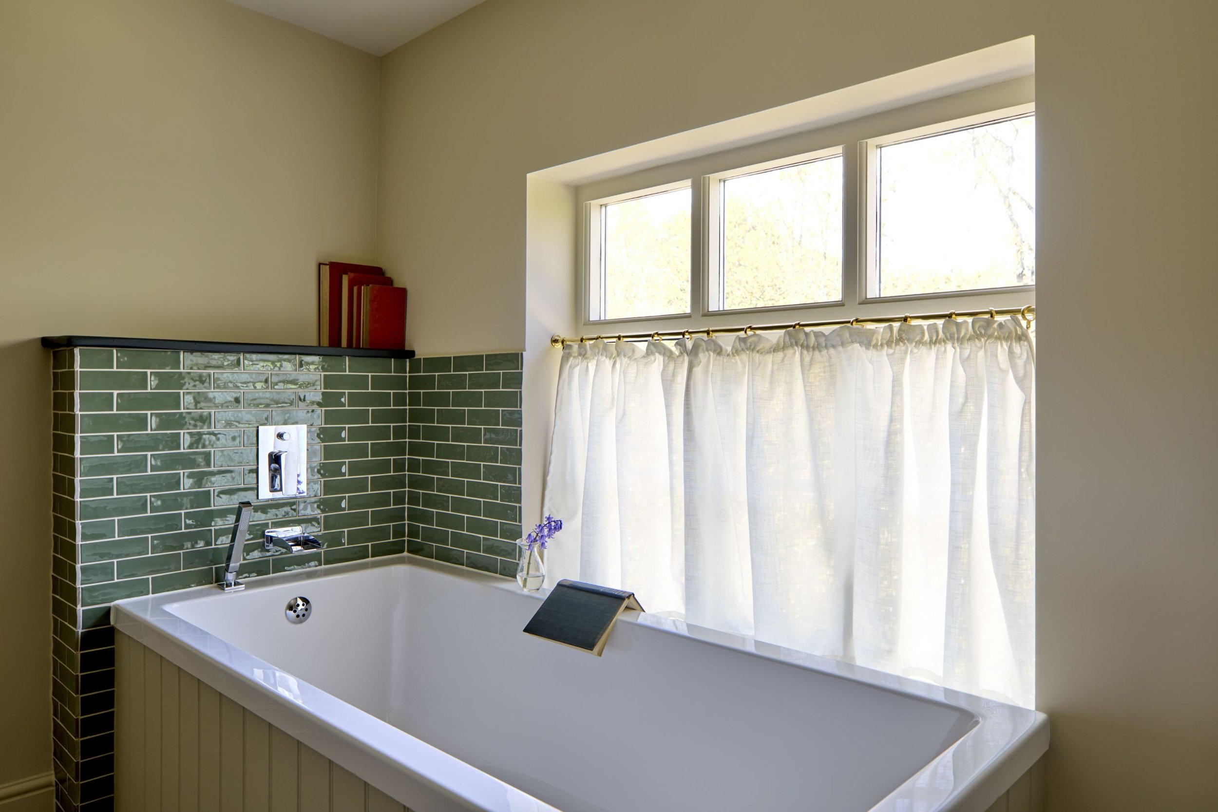 A bathroom with a white bathtub, green subway tile walls, a window with white curtains, and a small vase with purple flowers on the bathtub edge.