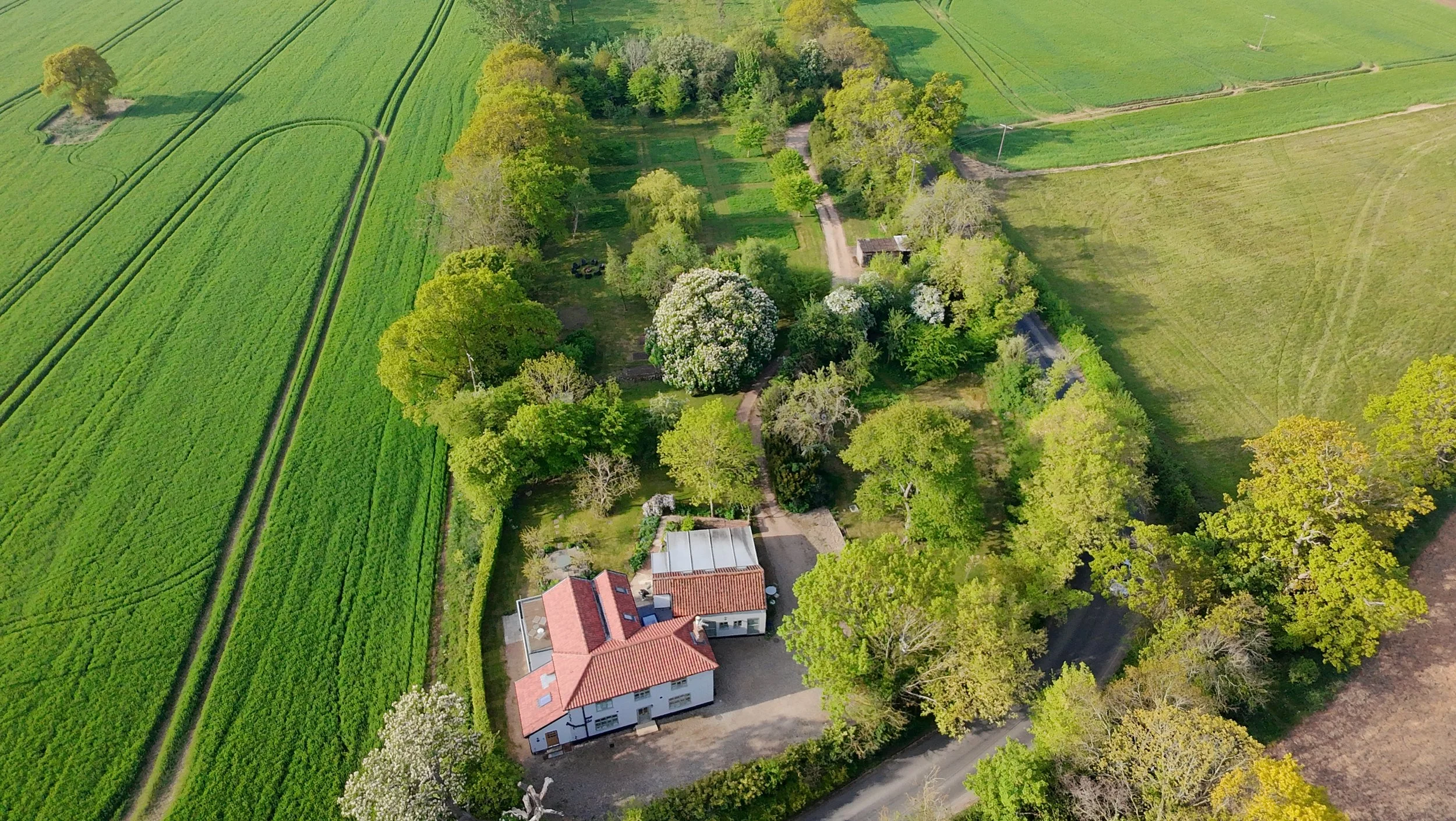 An aerial view of a rural property with multiple trees, a house with a red-tiled roof, and surrounding fields of green farmland.