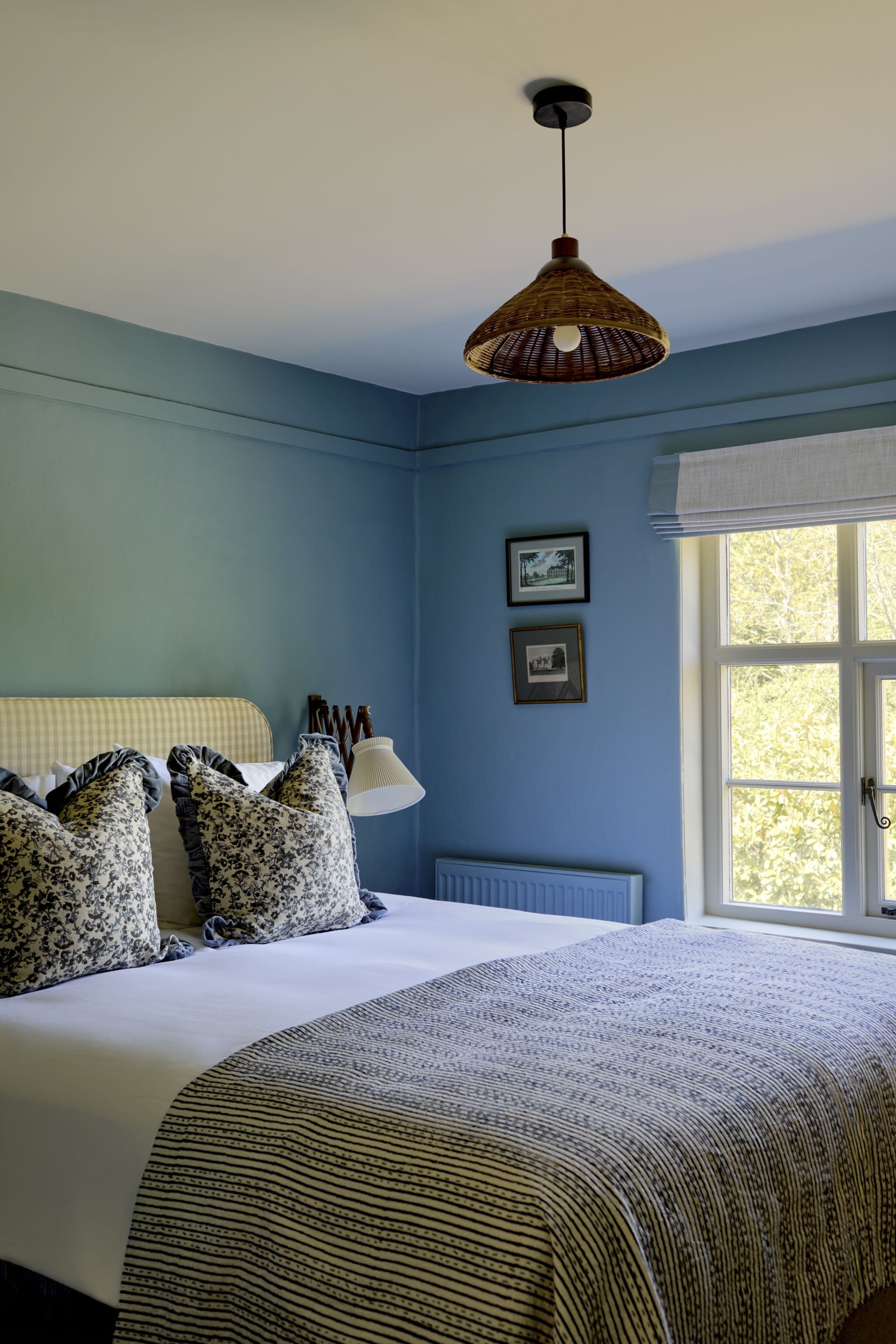 A bedroom with a bed, patterned pillows, a small bedside table with a lamp, a window with a view of trees, and a ceiling light with a woven basket shade.
