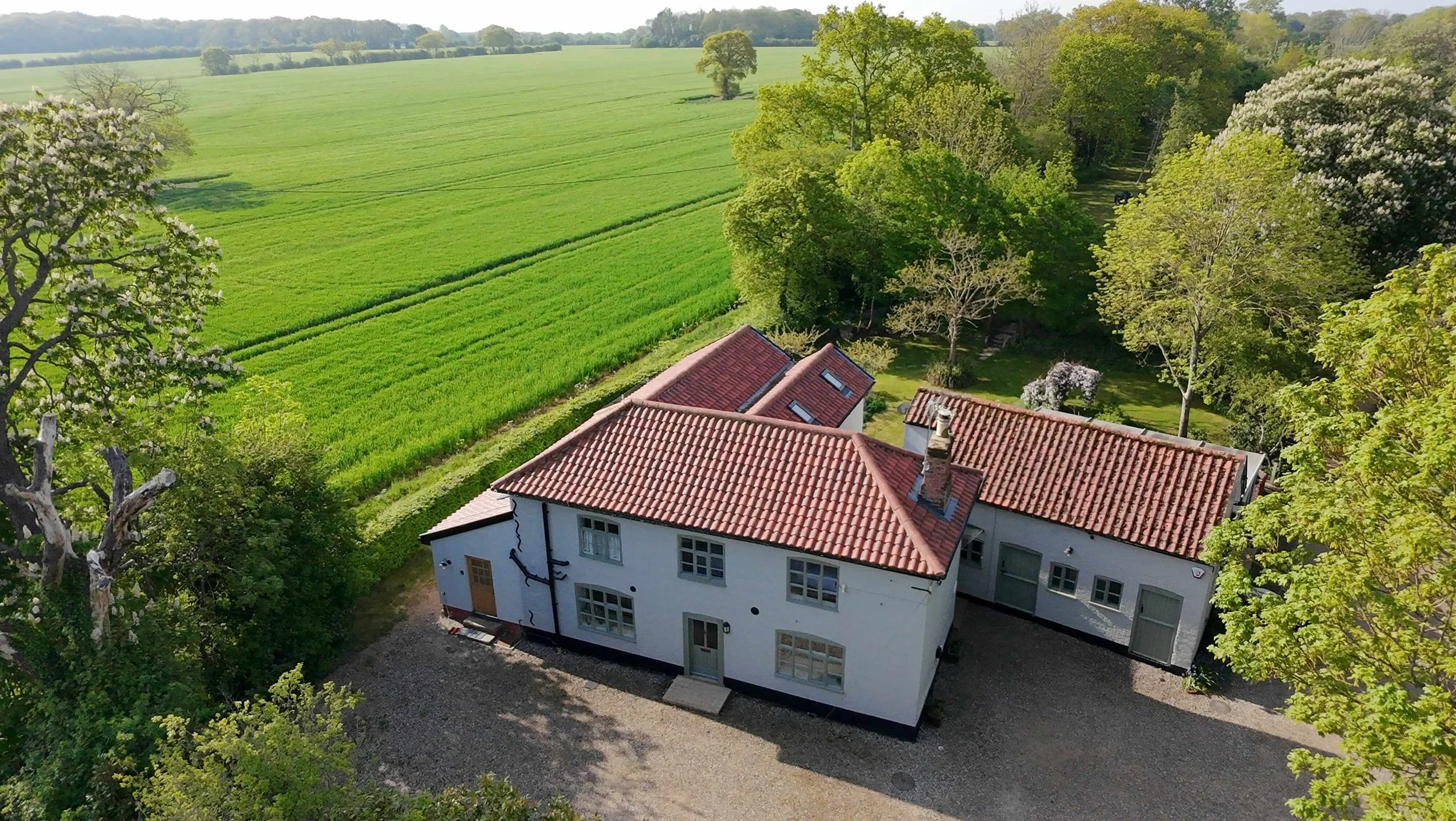 An aerial view of a house with a red-tiled roof, surrounded by lush green trees and open farmland with green fields extending into the horizon.