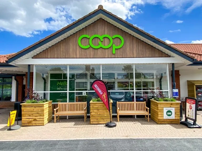 Exterior of a Coop store with a green sign, front window display, wooden benches, flower planters, and a red flag with a logo.