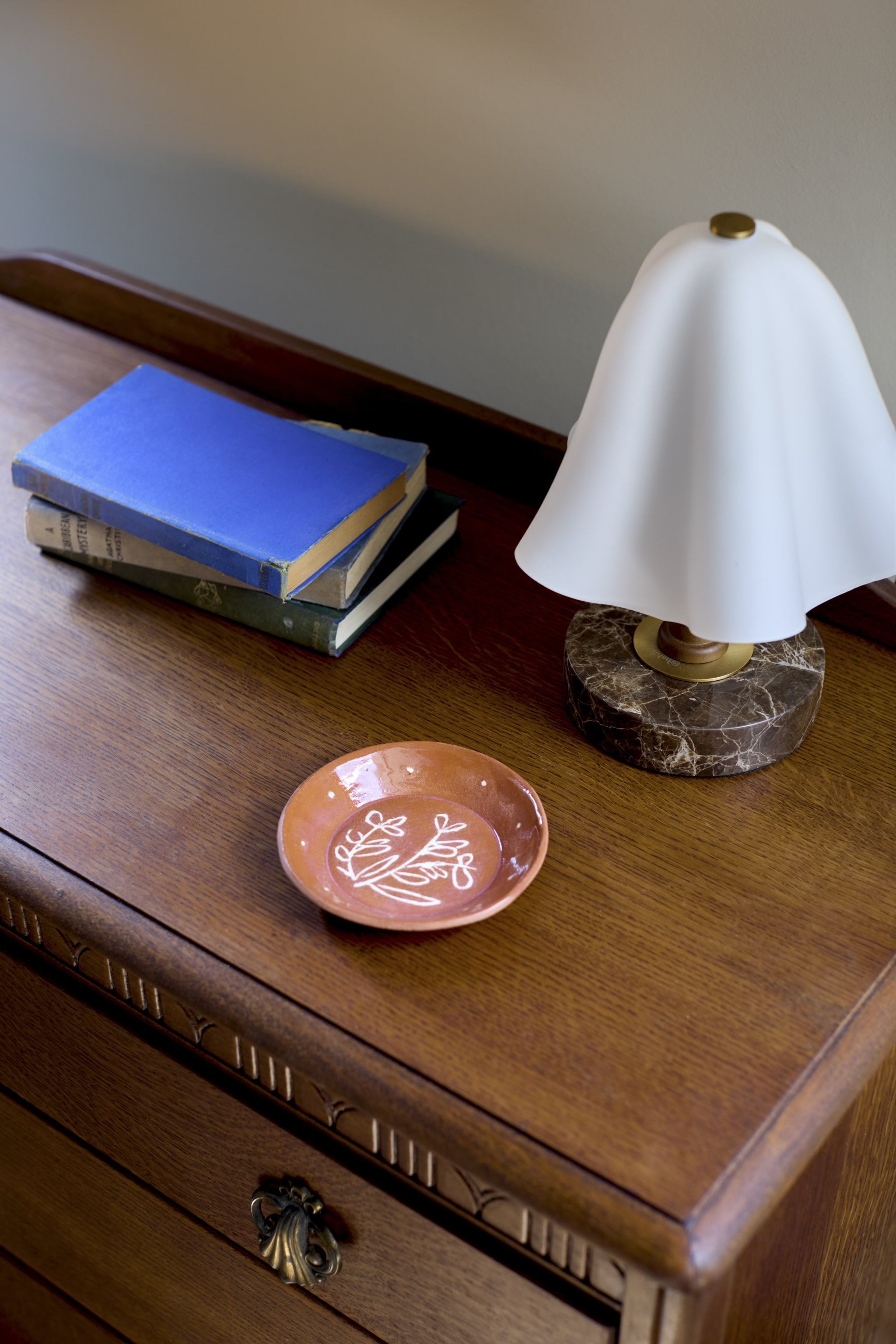A wooden table with a stack of three books, a small ceramic dish with a design, and a white Table lamp on a marble base. The table has carved edge details and a brass drawer handle.