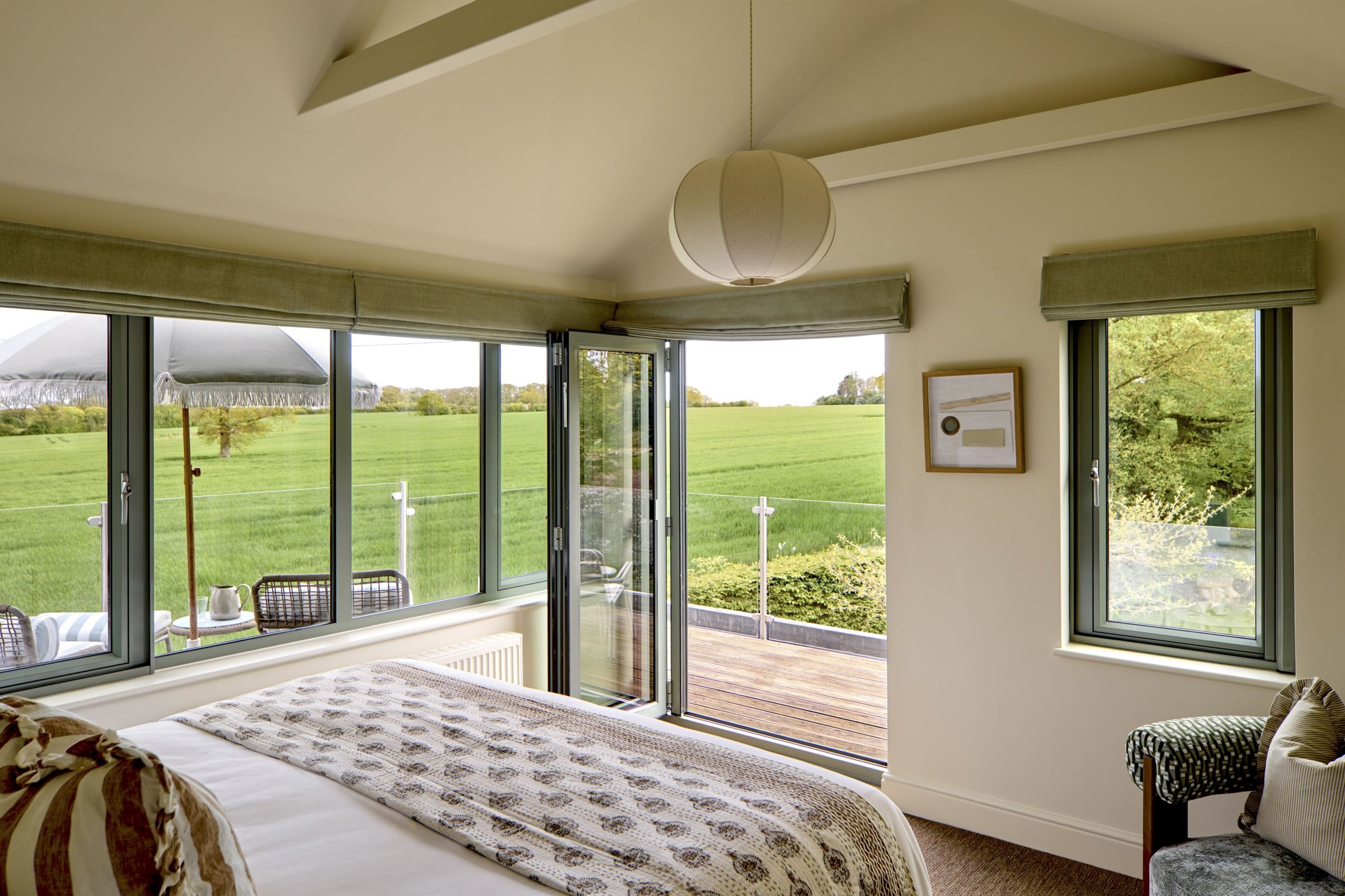 Interior view of a bedroom with large windows showing a green field outside, a bed with a patterned blanket, a framed artwork on the wall, and a sliding glass door leading to a wooden deck.