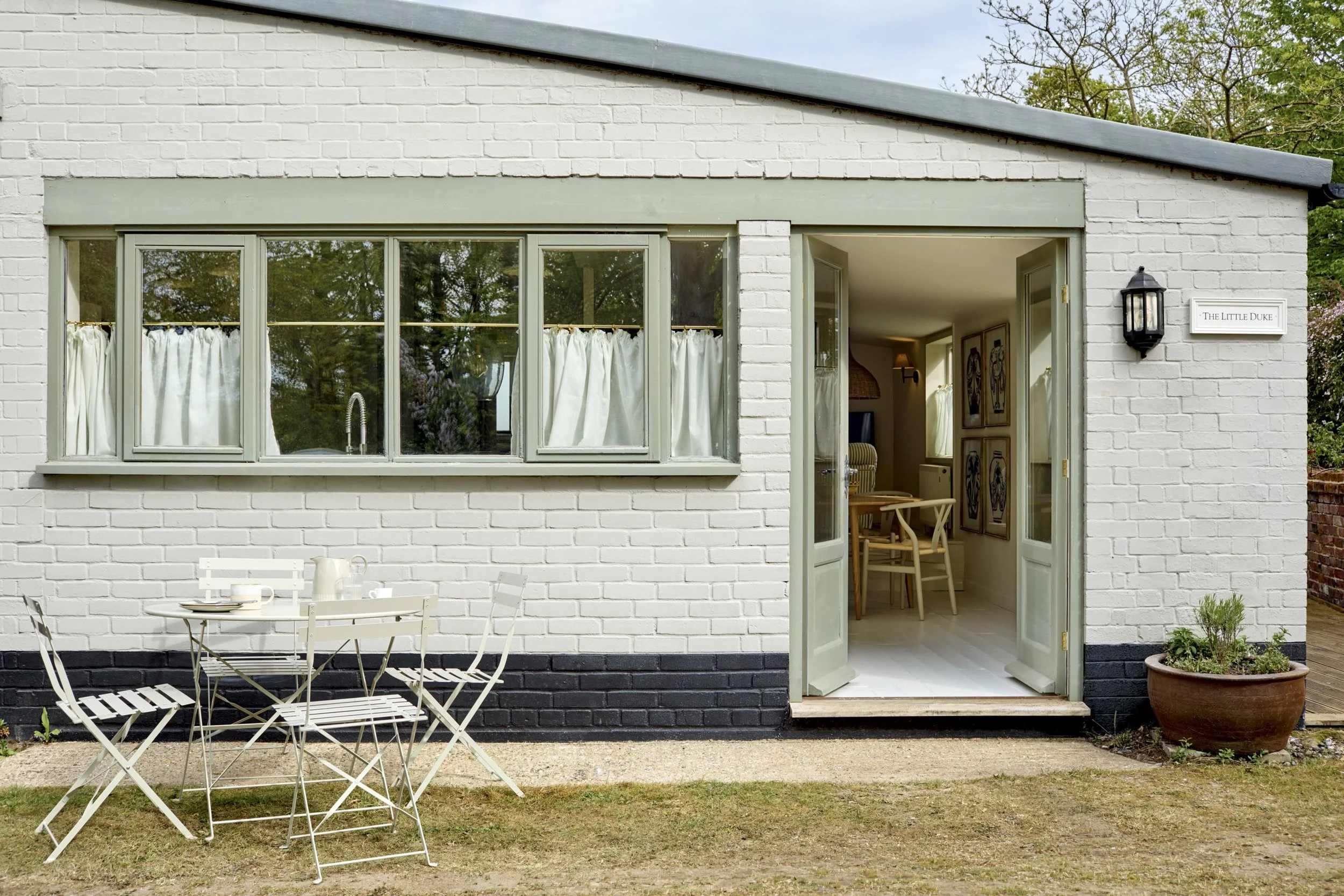 A house with white brick exterior, large windows, and a door open to a cozy interior. Outside, there is a white patio table with chairs and a large potted plant on the ground.