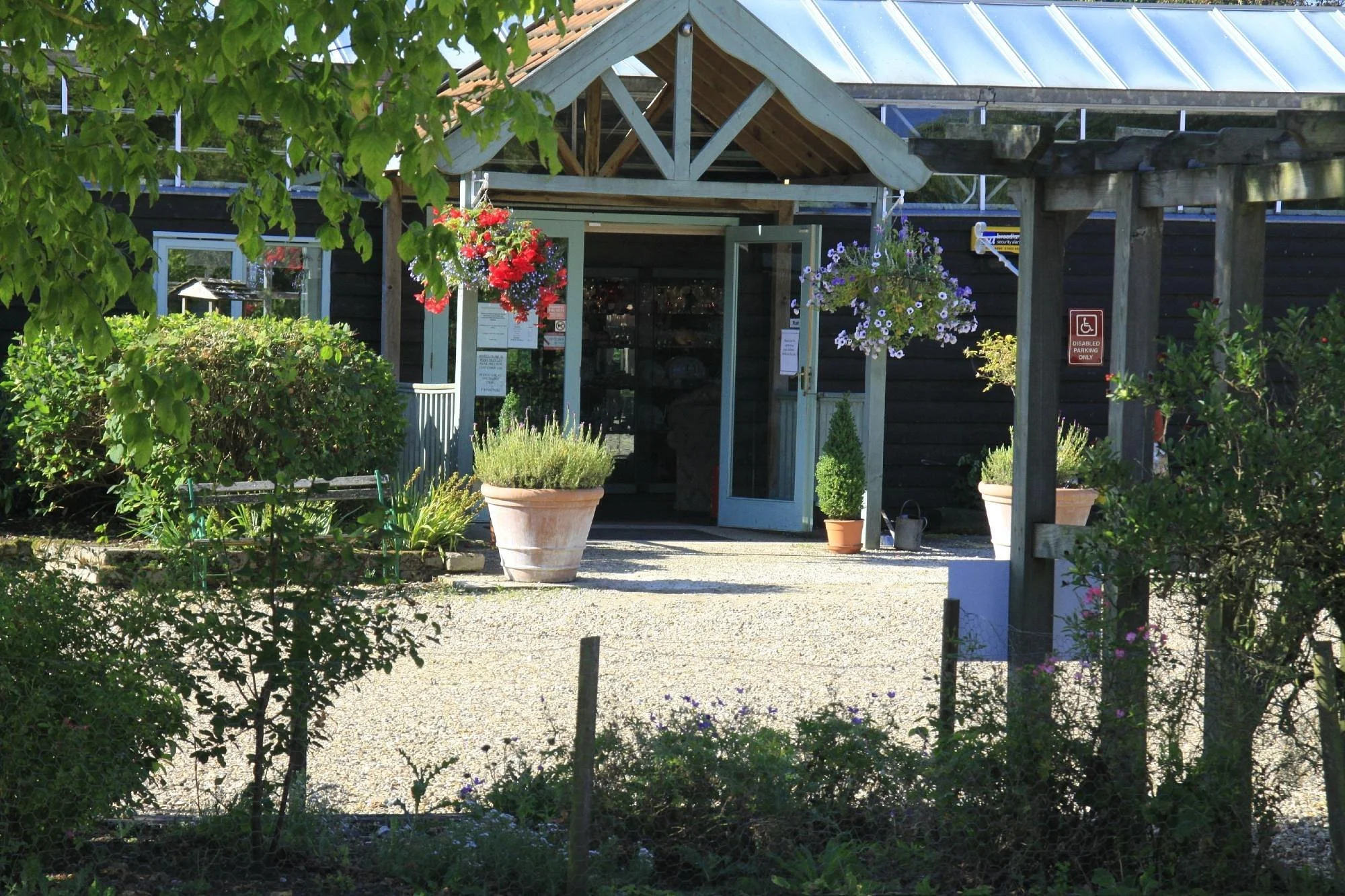 Entrance of a rustic shop or café with an open glass door, surrounded by greenery, hanging and potted flowers, and a gravel pathway. There is a red and white sign indicating accessible parking.