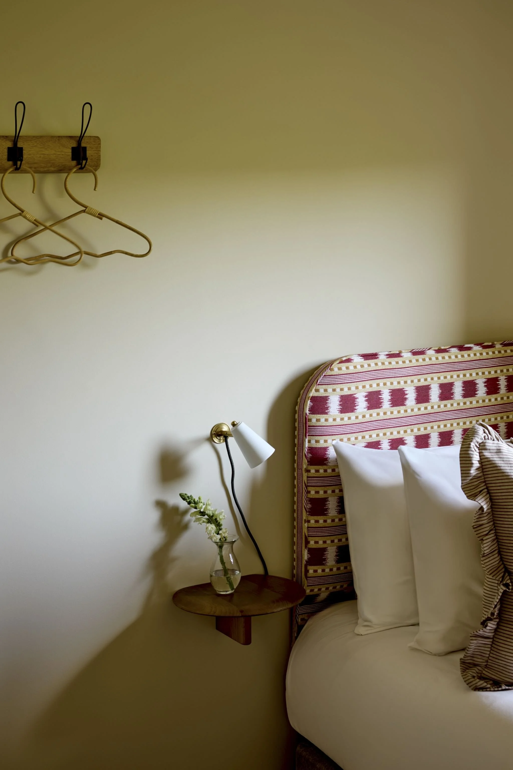 A bedroom corner with a bed featuring a red and white striped headboard, a white pillow, a small wooden side table, a white flower in a glass vase, a white wall lamp, a wall-mounted wooden coat rack with two empty bamboo hangers, and a beige wall.
