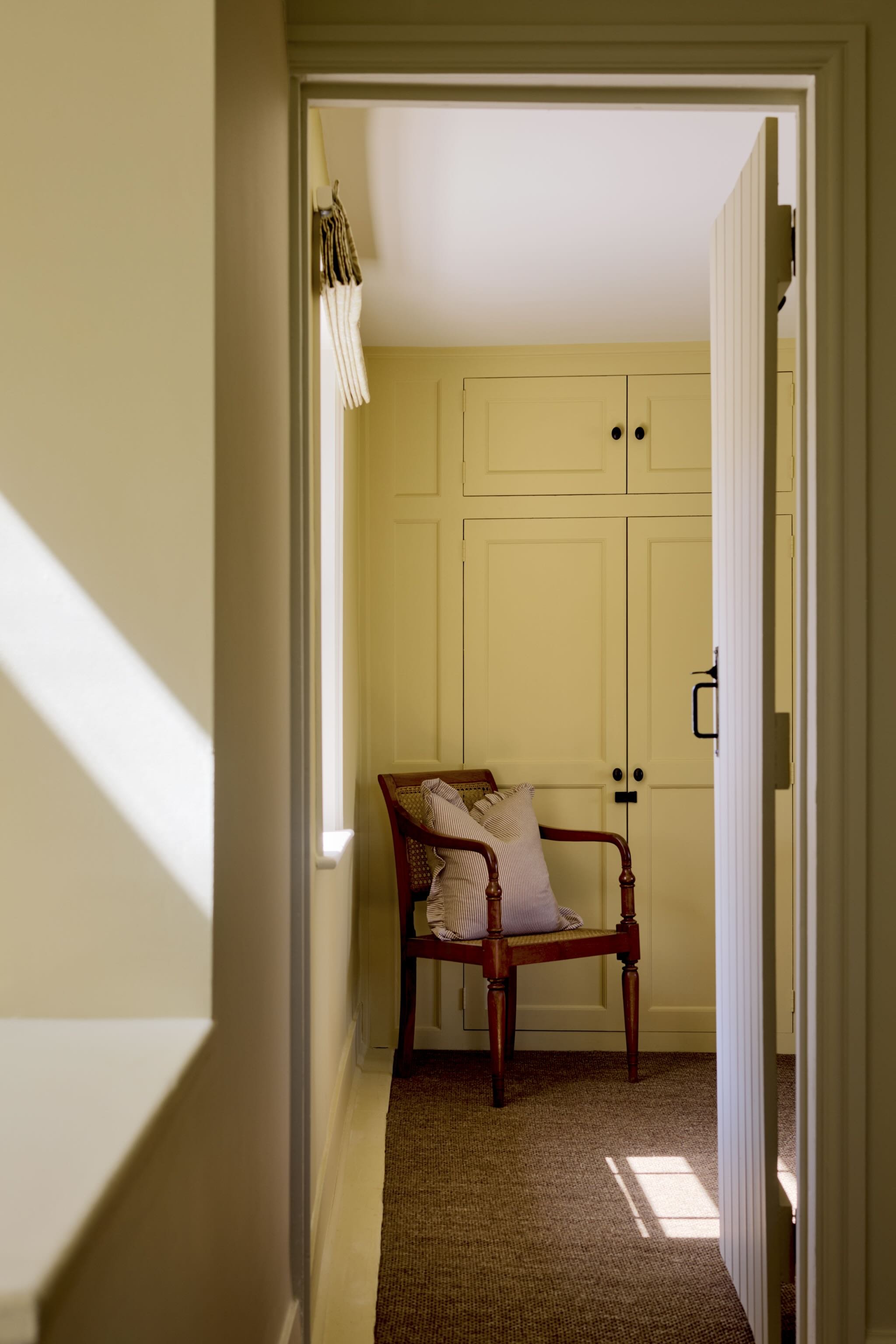 A yellow-painted hallway leading to a small sitting area with a wooden chair that has a striped pillow and cushion, against a backdrop of yellow cabinetry and sunlight coming through a window.