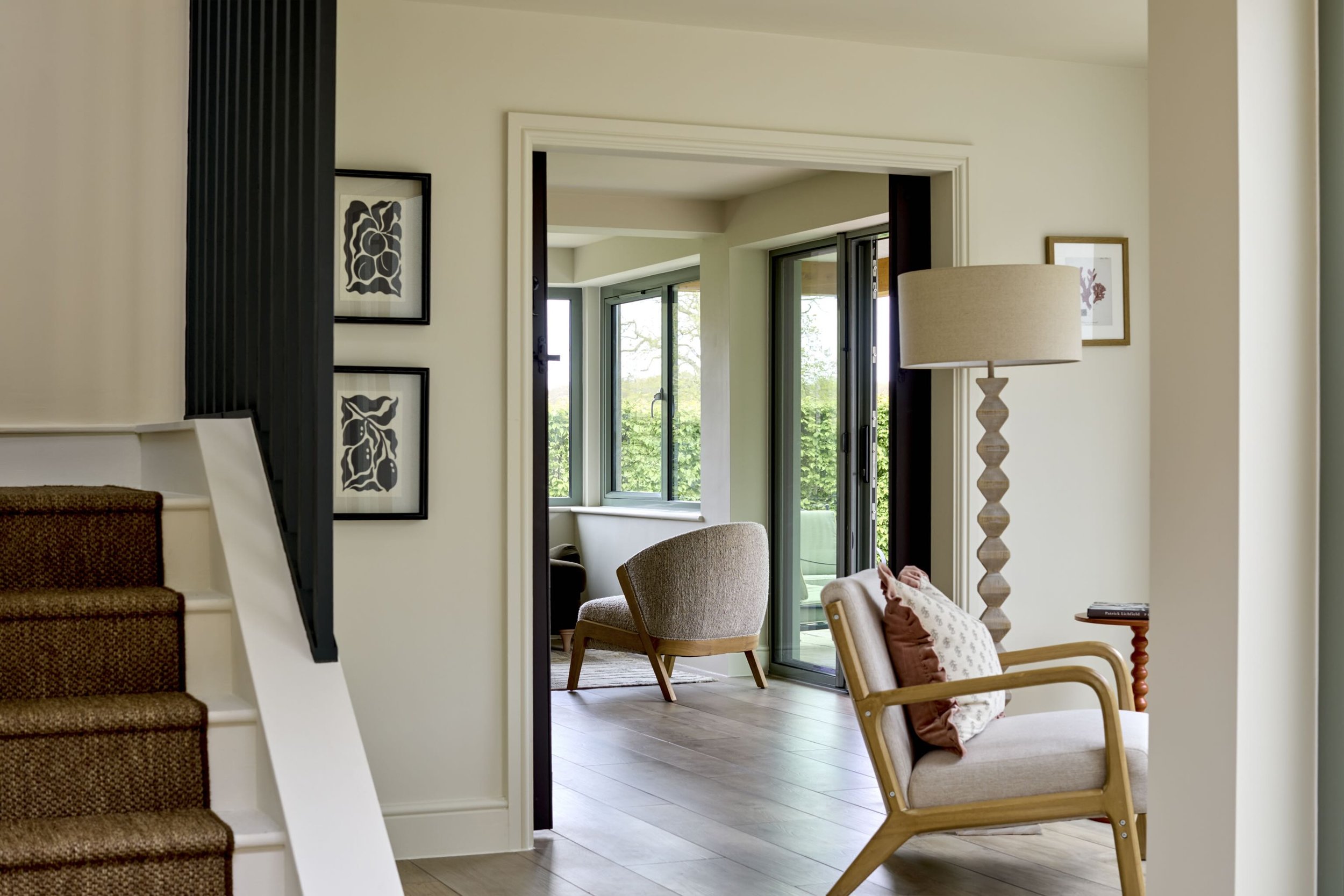 Living room with armchairs, a tall wooden lamp, framed pictures on the wall, and an open sliding door leading to a balcony with greenery outside.