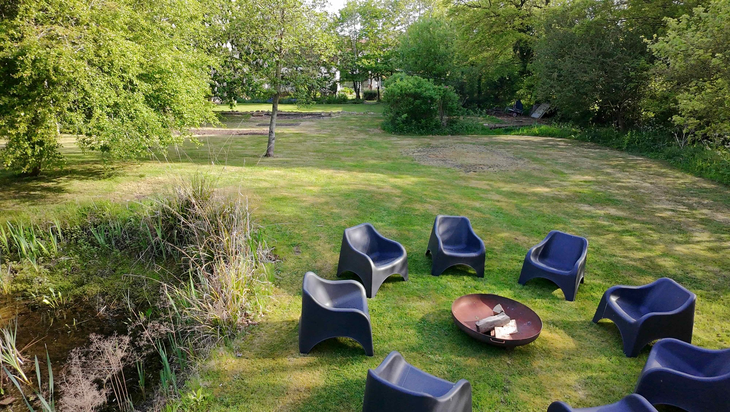 A backyard with a grassy lawn, surrounded by trees and bushes. There are five blue plastic chairs around a fire pit with some wood inside. The background shows more trees and a patio area.