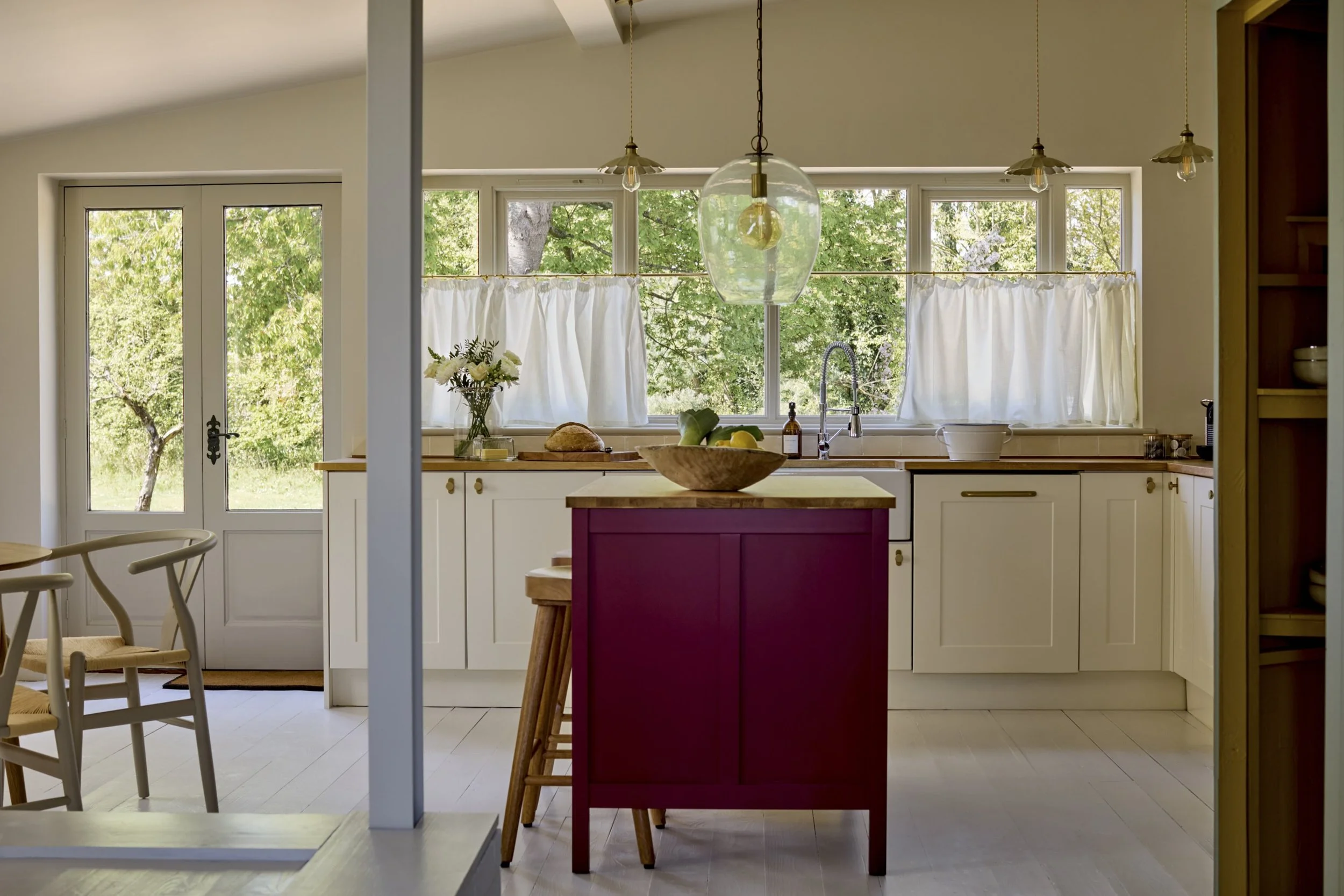 View of a kitchen with large windows showing green trees outside, a red kitchen island, white cabinets, and pendant lights.