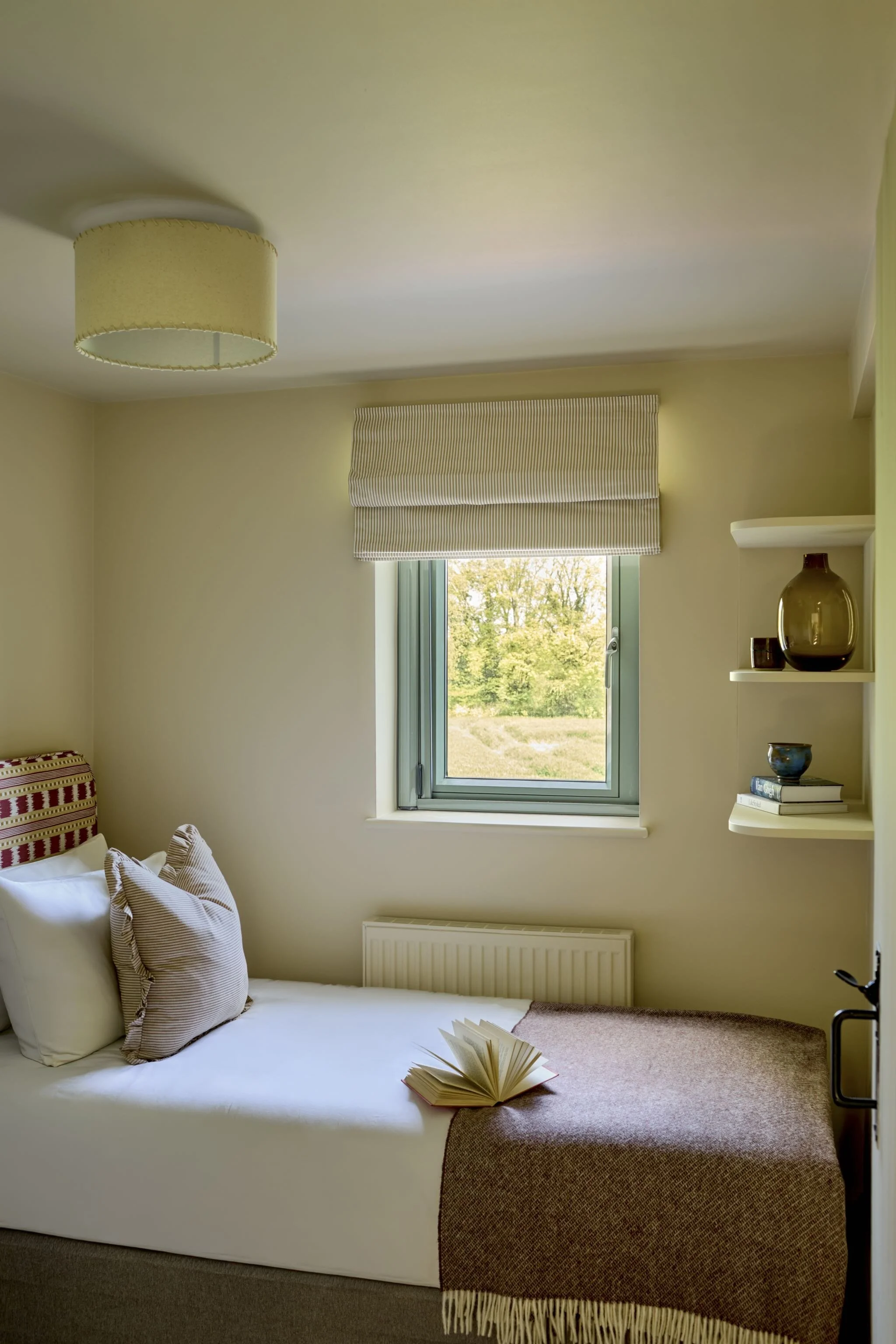 A cozy bedroom corner showing a bed with striped pillows, a window with a beige Roman shade, a shelf with a brown vase, an open book on the bed, and a view of trees outside the window.