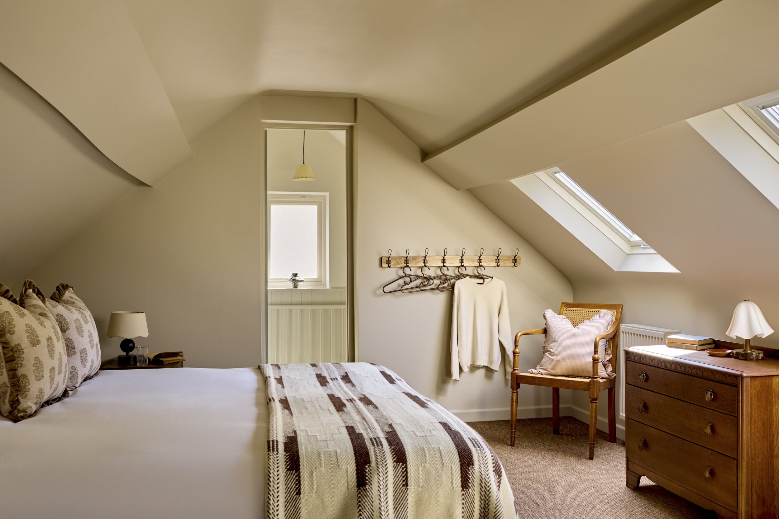 A cozy bedroom with a sloped ceiling, beige walls, and natural light coming from skylights. The bed has patterned pillows and a blanket, with a nightstand and lamps. A wooden chair with a cushion and a dresser with books are also visible.