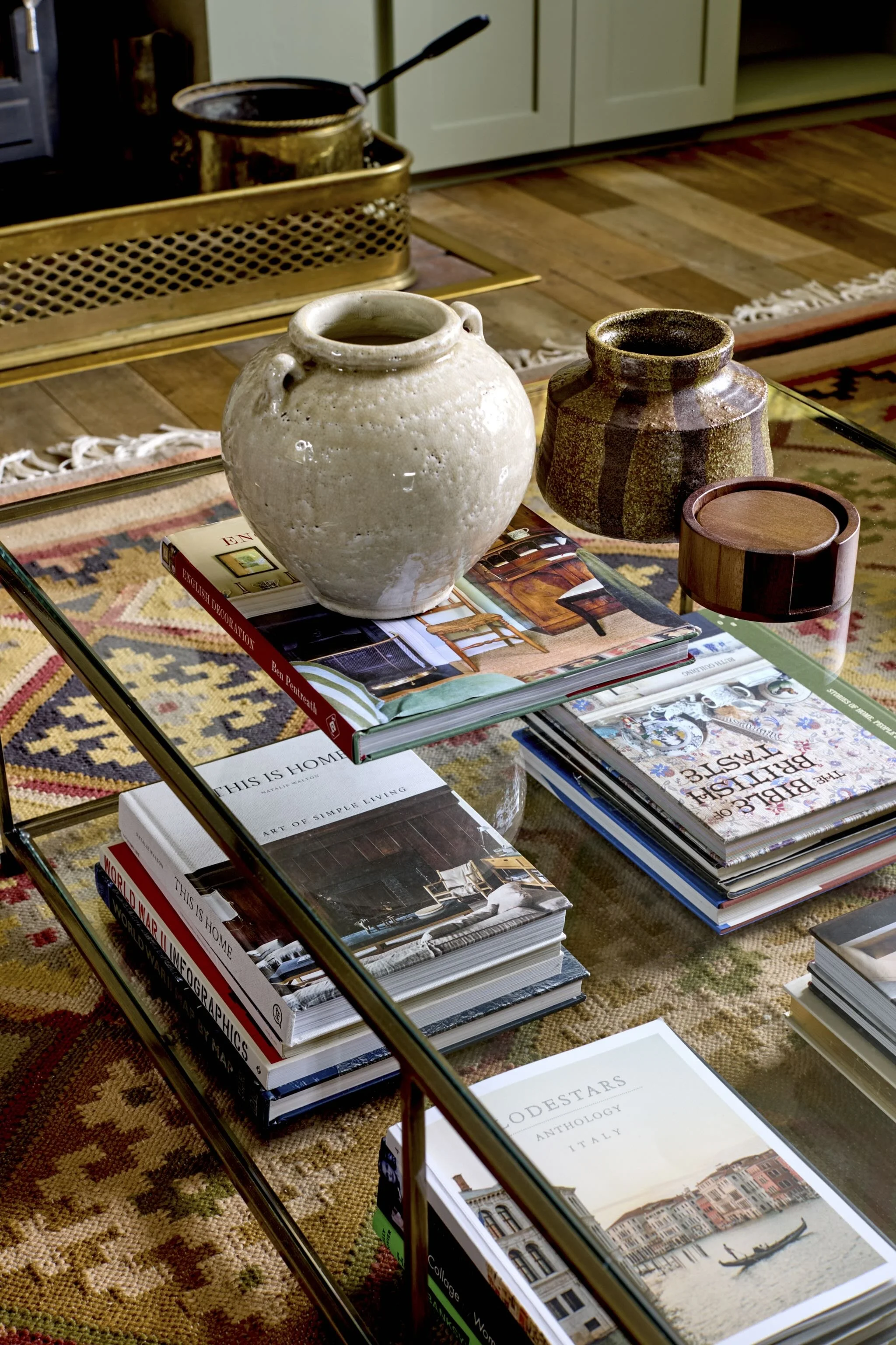 A glass coffee table with art and interior design books, ceramic vases, and a small wooden round container on top, situated on a patterned rug in a cozy living room.