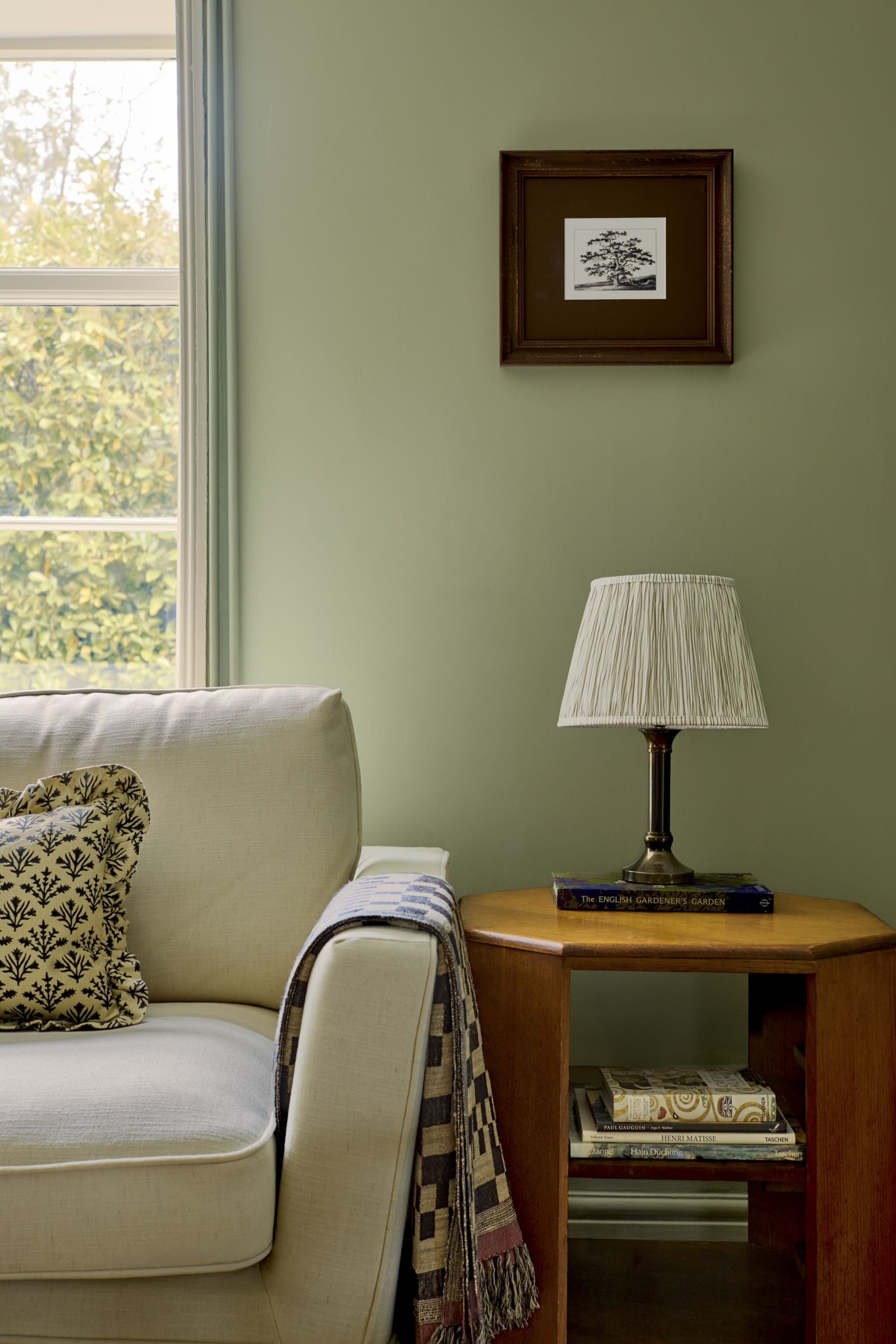 Living room corner with cream sofa, matching chair, throw pillow, wooden side table holding a lamp and art books, green wall with framed black and white tree picture, window with greenery outside.