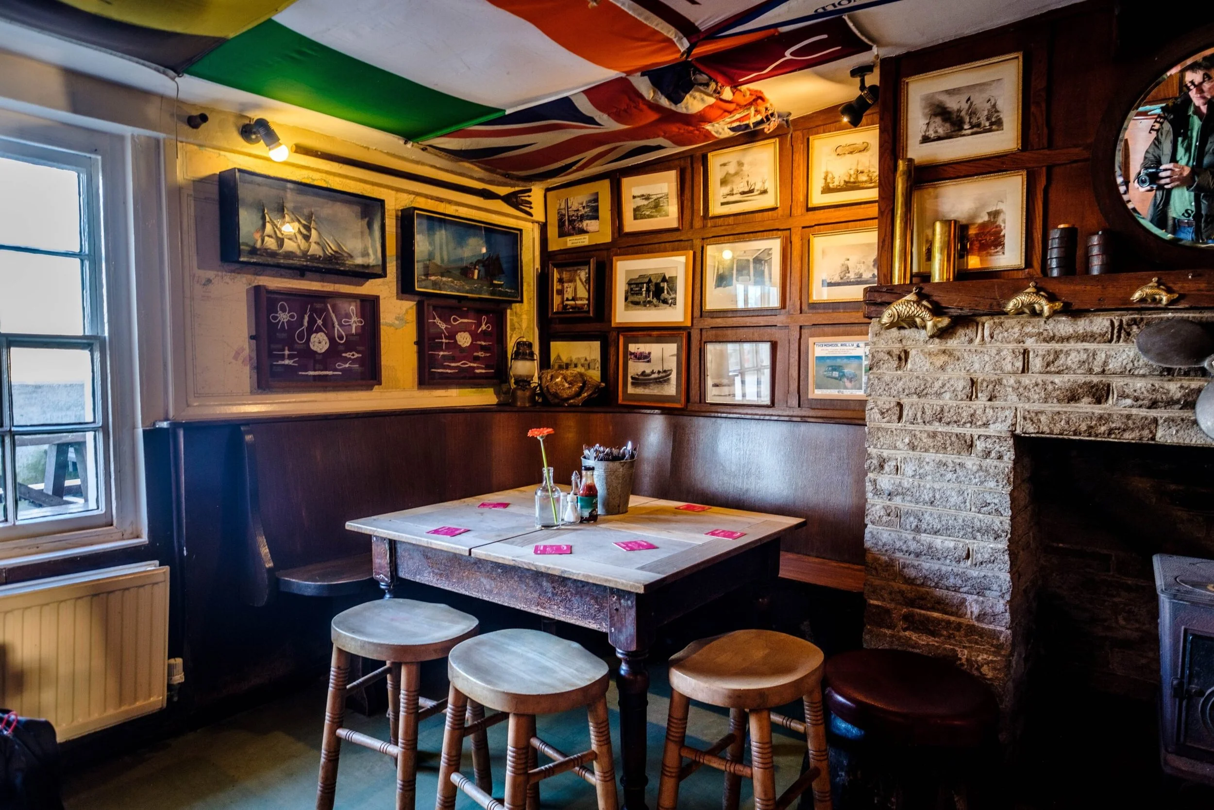 Cozy restaurant corner with wooden paneling, framed maritime paintings, a brick fireplace decorated with golden fish figurines, and a table set with pink placemats, jars, and utensils near a window.