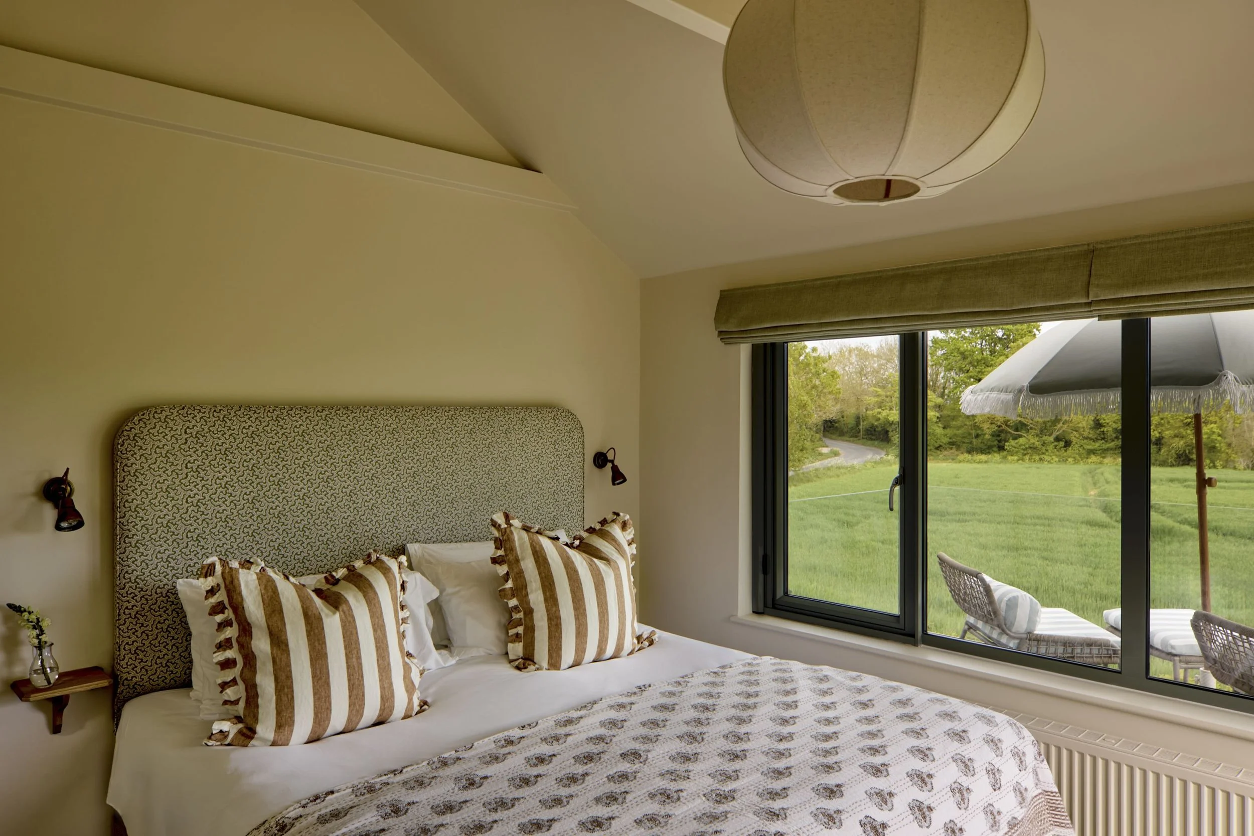 A bedroom with a window overlooking a green landscape, a bed with a patterned headboard and striped pillows, and a ceiling light fixture.