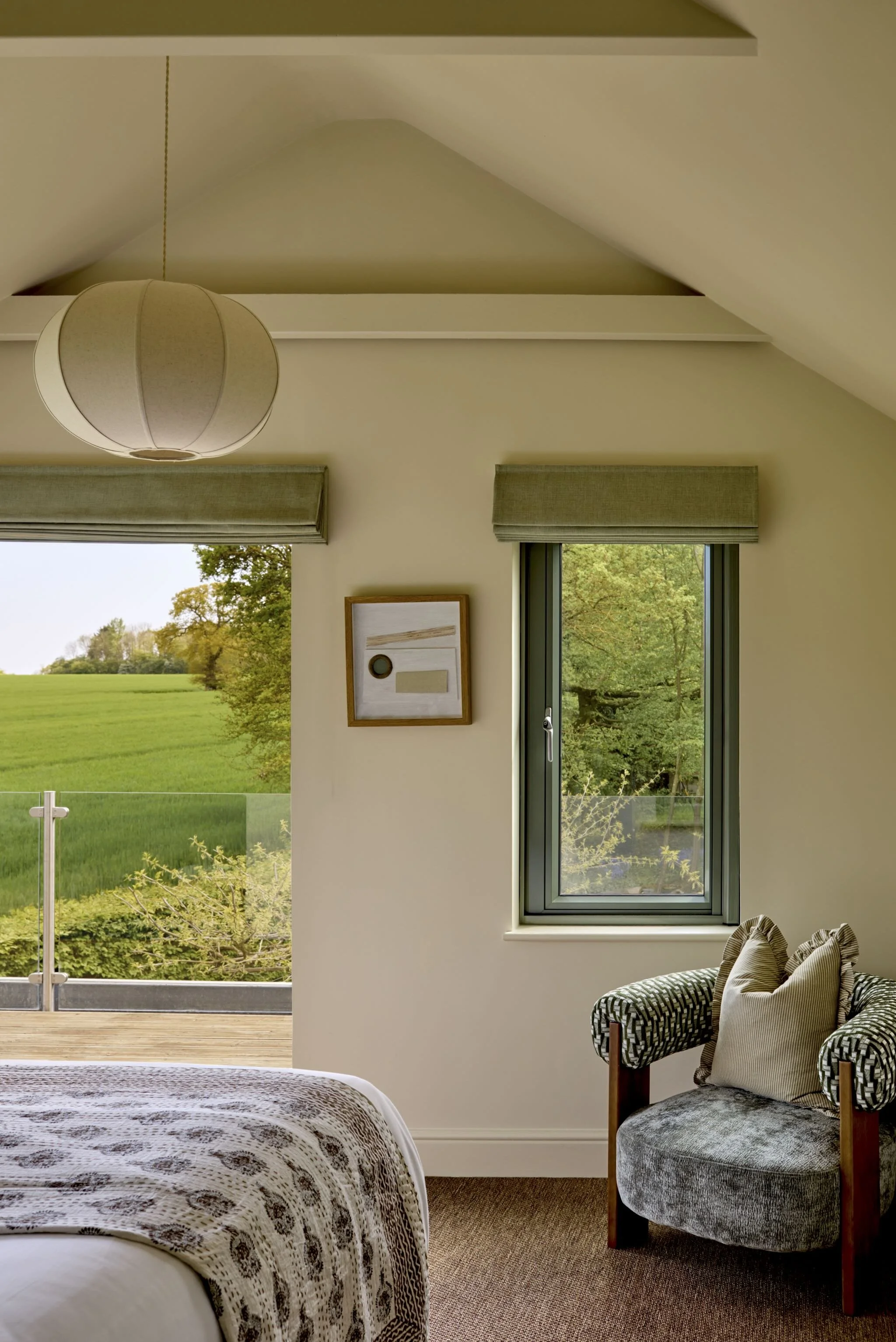A bedroom with a large window showing green trees and grass outside, a beige armchair with a patterned cushion, and a bed with a patterned blanket. The room has a ceiling light, framed artwork on the wall, and gray window blinds.