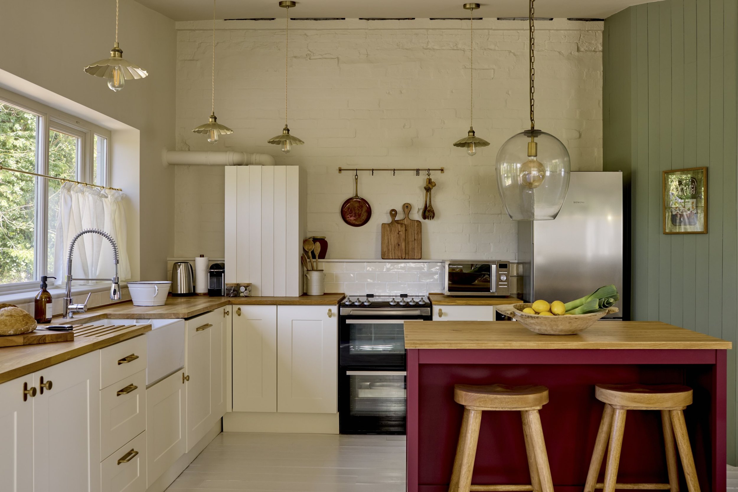 A cozy kitchen with white cabinets, a wooden countertop, and a green accent wall. Features include a window with white curtains, a kitchen sink with a curved faucet, pendant lights, and a kitchen island with a bowl of lemons and leeks. Wooden stools 
