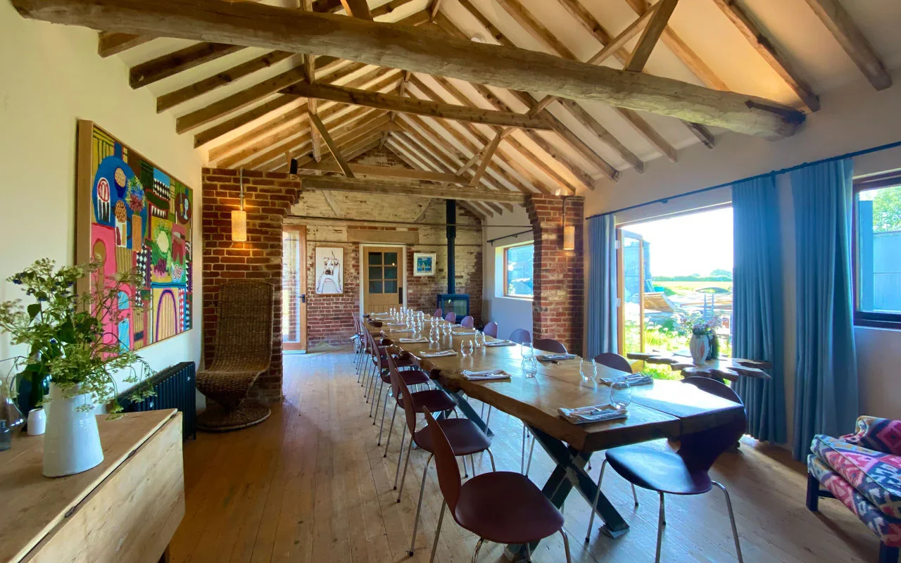 Interior of a rustic dining room with a long wooden table set for a meal, surrounded by chairs. The room features exposed wooden beams, brick walls, large windows with blue curtains, and outdoor scenery visible through the windows.
