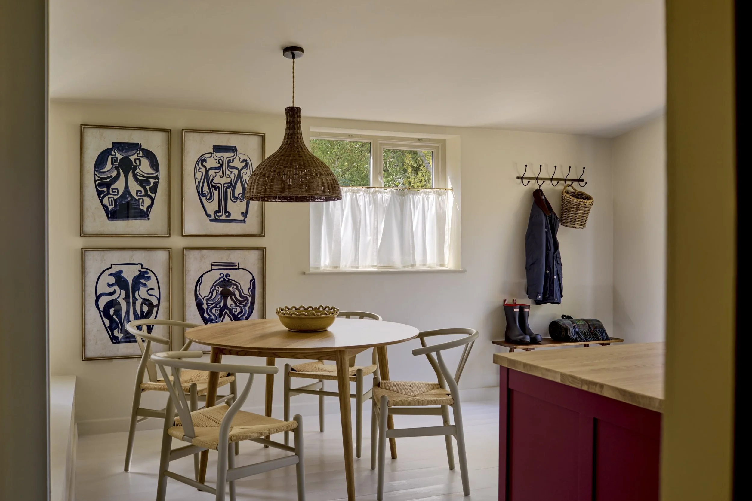A cozy kitchen corner with a wooden dining table and chairs, wall art featuring blue vase illustrations, a window with white curtains, a woven pendant light, and a coat rack with a jacket, a woven basket, and rain boots.
