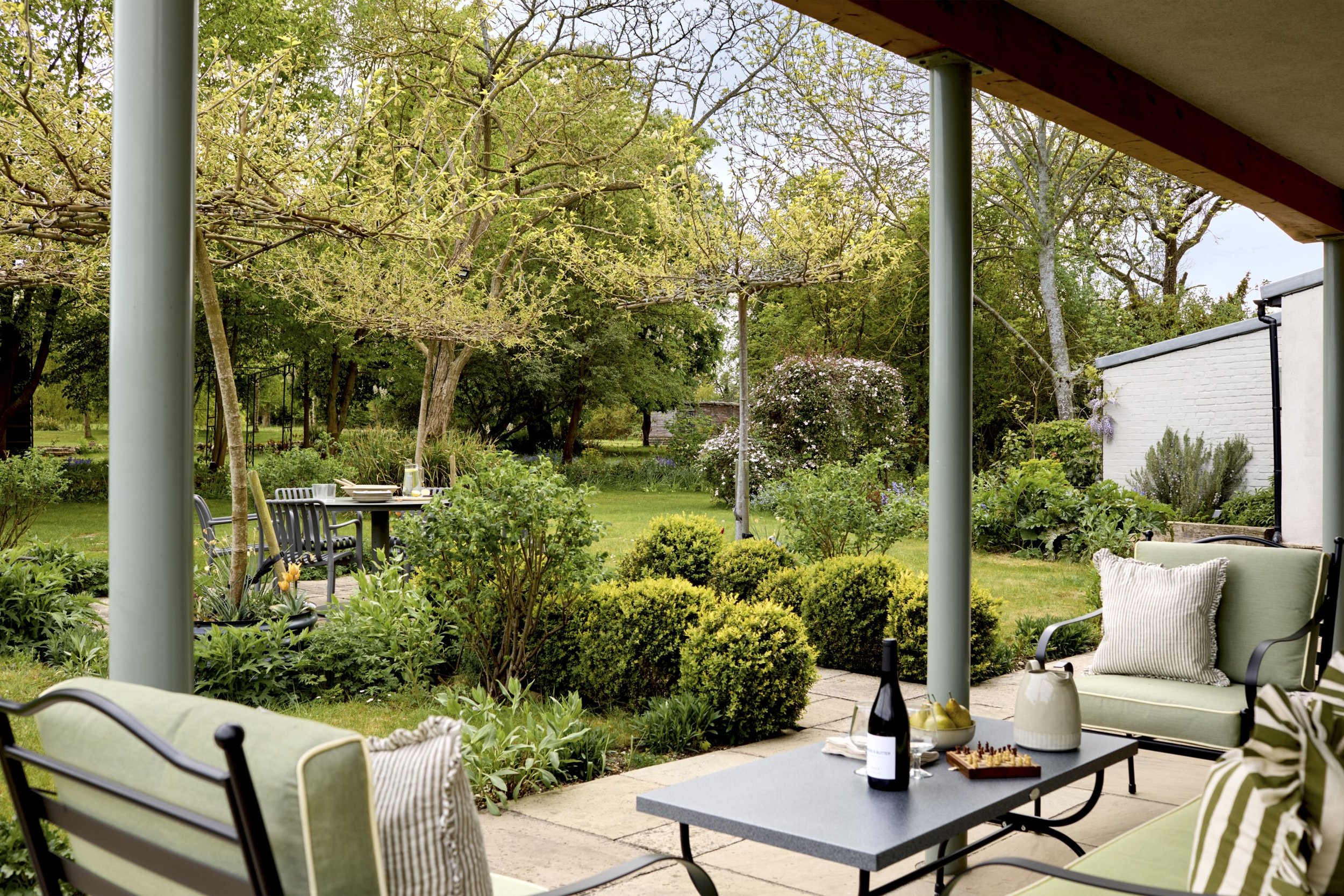 A cozy outdoor patio with green cushioned furniture, a table with a bottle of wine, and lush greenery in the background with trees and flowering bushes.