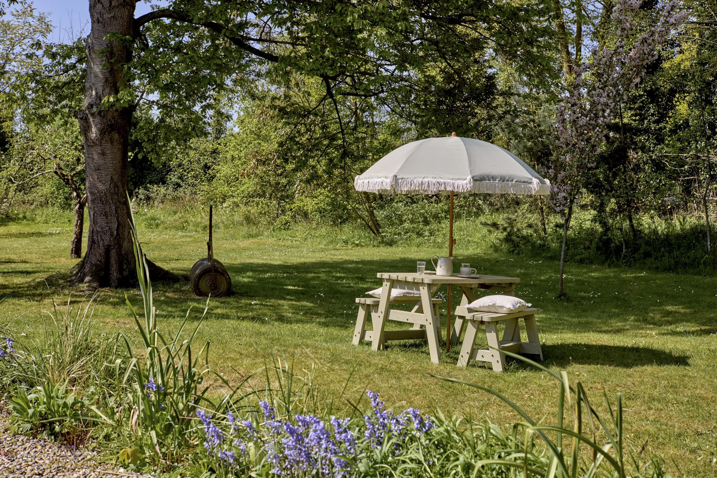 A peaceful outdoor garden scene with a large tree and lush green foliage. There is a white umbrella over a wooden table set with a teapot, cups, and glasses. Purple flowers bloom in the foreground.