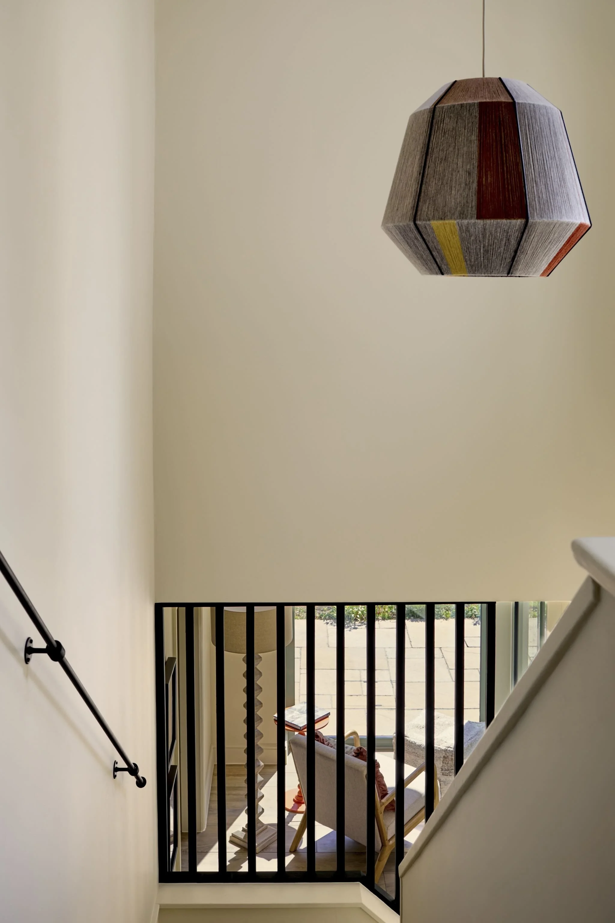 Interior view of a second-floor balcony with a black metal railing, a beige armchair, a small side table, a table lamp with a beige shade, and an open view to a patio with outdoor seating and greenery.