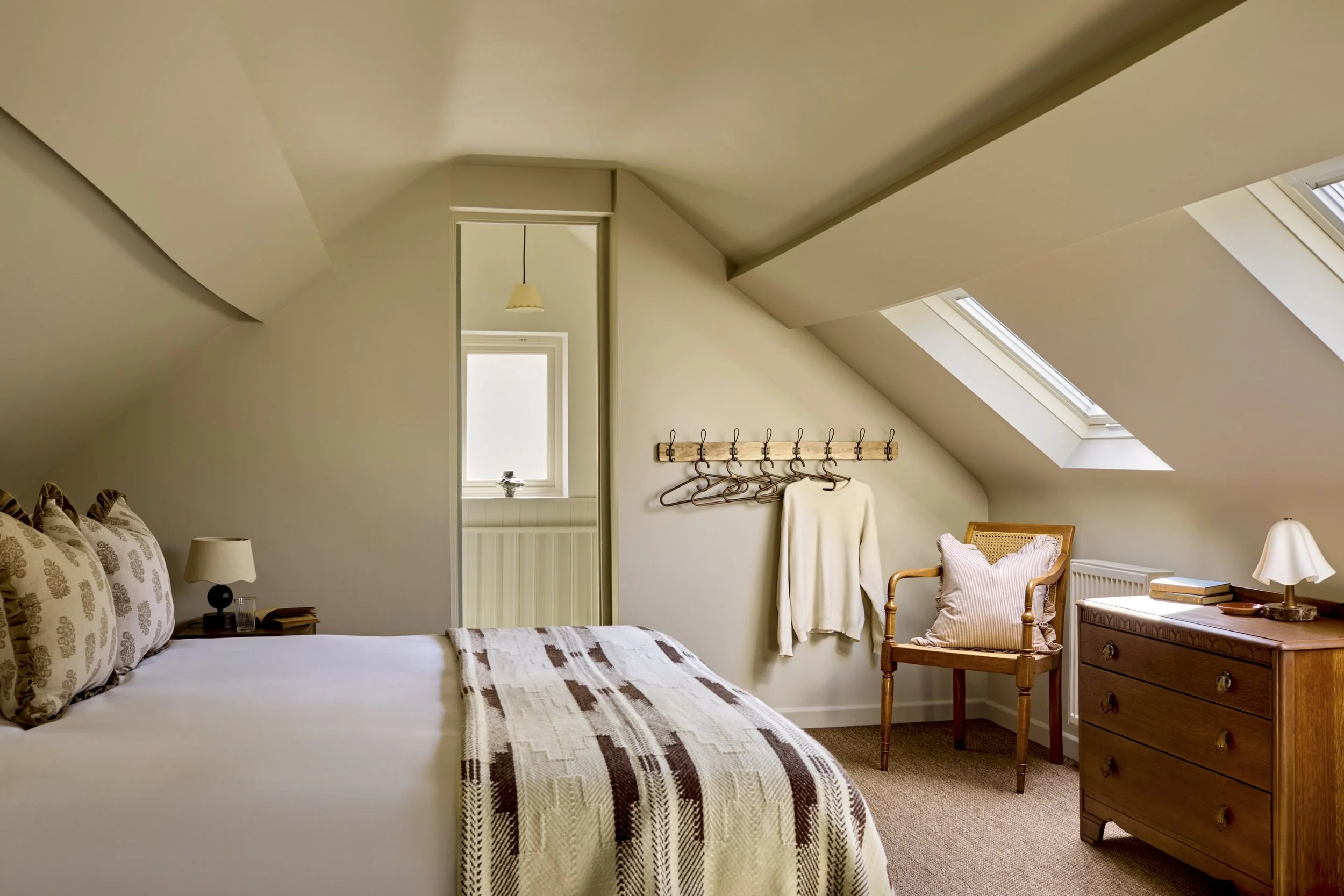 An attic bedroom with a sloped ceiling, skylights, and a window, decorated with a bed, wooden dresser, chair, and wall hooks with hanging clothes.