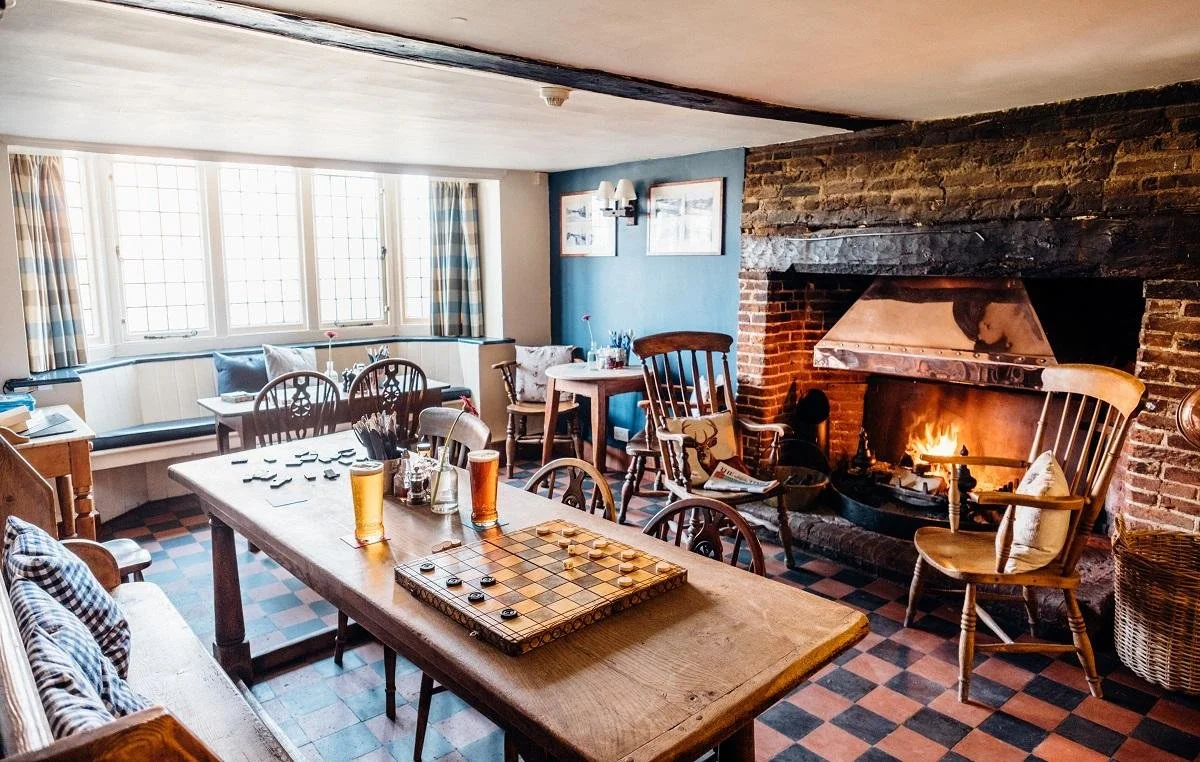 Cozy living room with a brick fireplace and a game of checkers on the table, surrounded by wooden chairs, a window seat with cushions, and warm lighting.