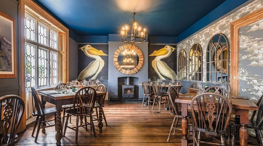 A cozy dining room with dark blue walls and wooden floors, decorated with large pelican murals, a round mirror, and a wood-burning stove. The room features wooden tables and chairs, with a chandelier hanging from the ceiling.