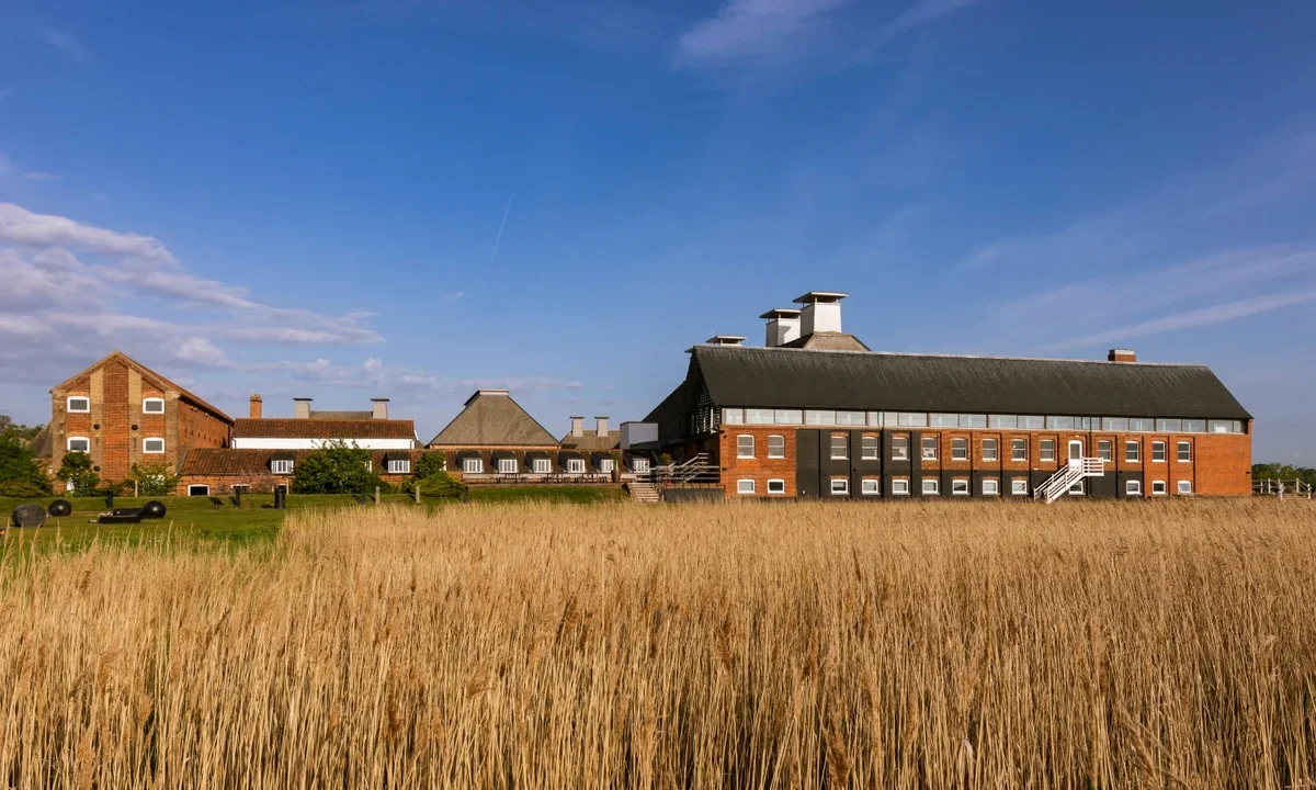 A large brick building with a black roof on a grassy field with tall grass, under a clear blue sky with a few clouds.
