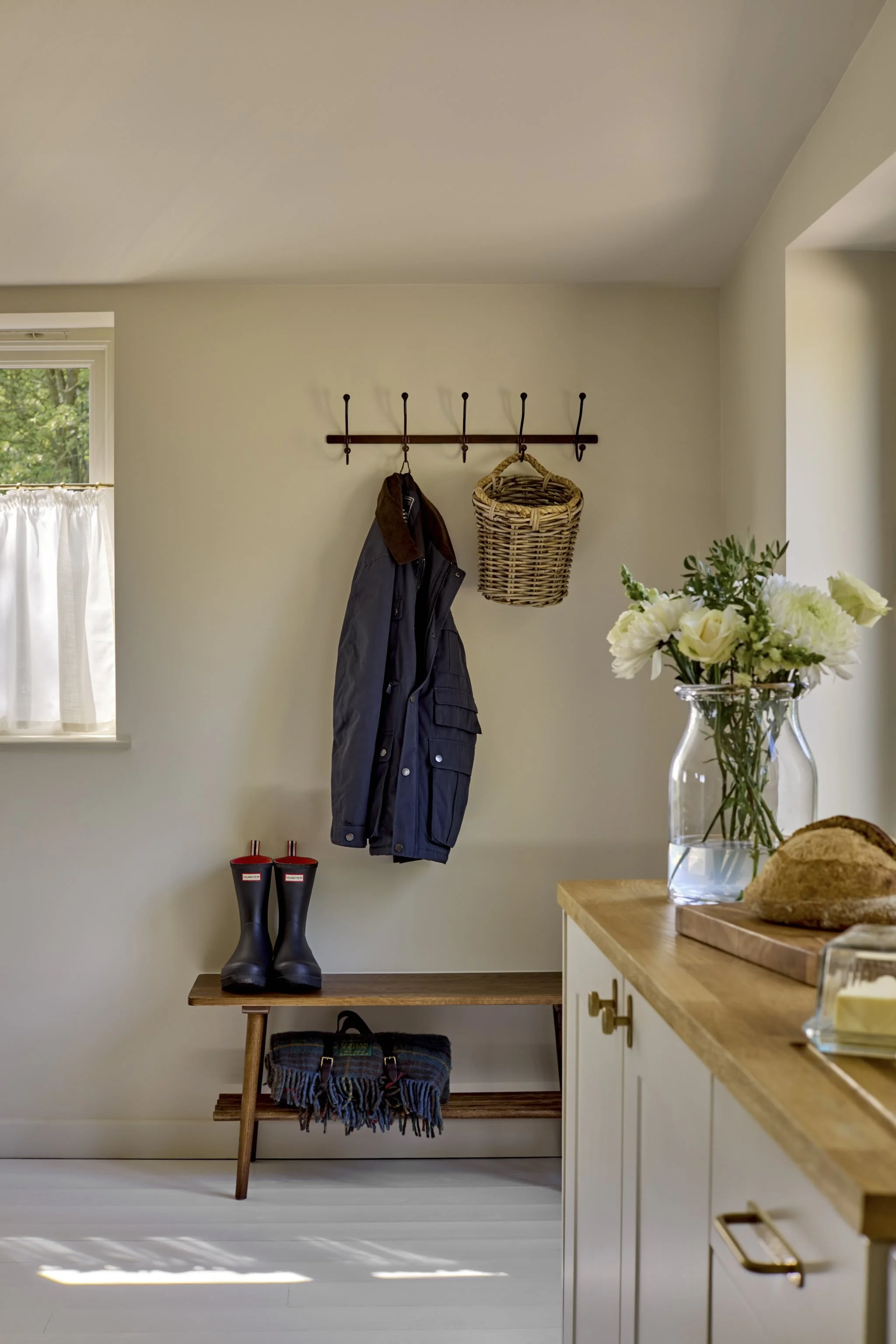 Living room entryway with a wall-mounted coat rack holding a jacket and a woven basket, a pair of rubber boots on a wooden bench, a window with white curtains, a vase of white flowers on a kitchen counter, and sunlight casting shadows on the floor.