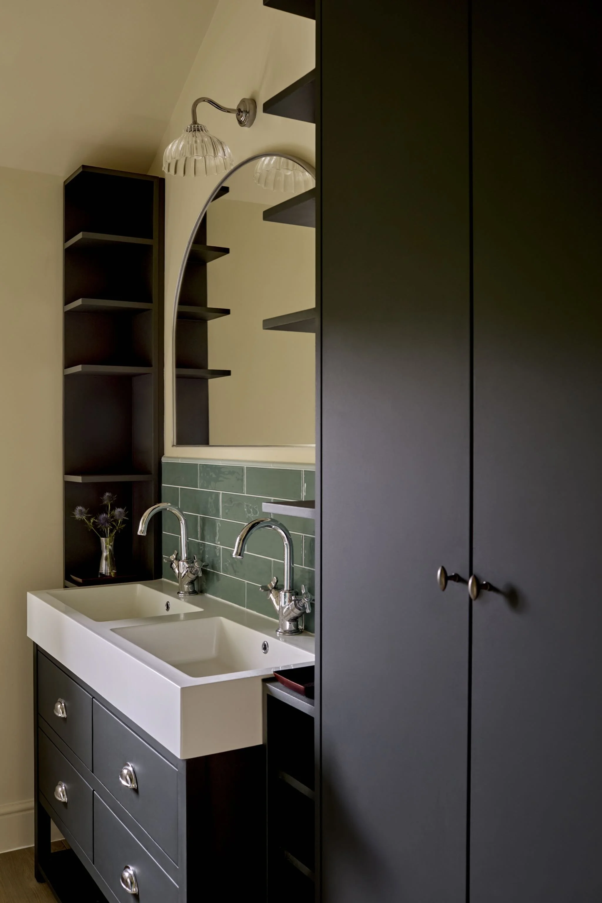 A kitchen with a white double sink, green tiled backsplash, dark cabinets, and a black shelving unit. There is a mirror reflecting a wall and a light fixture.