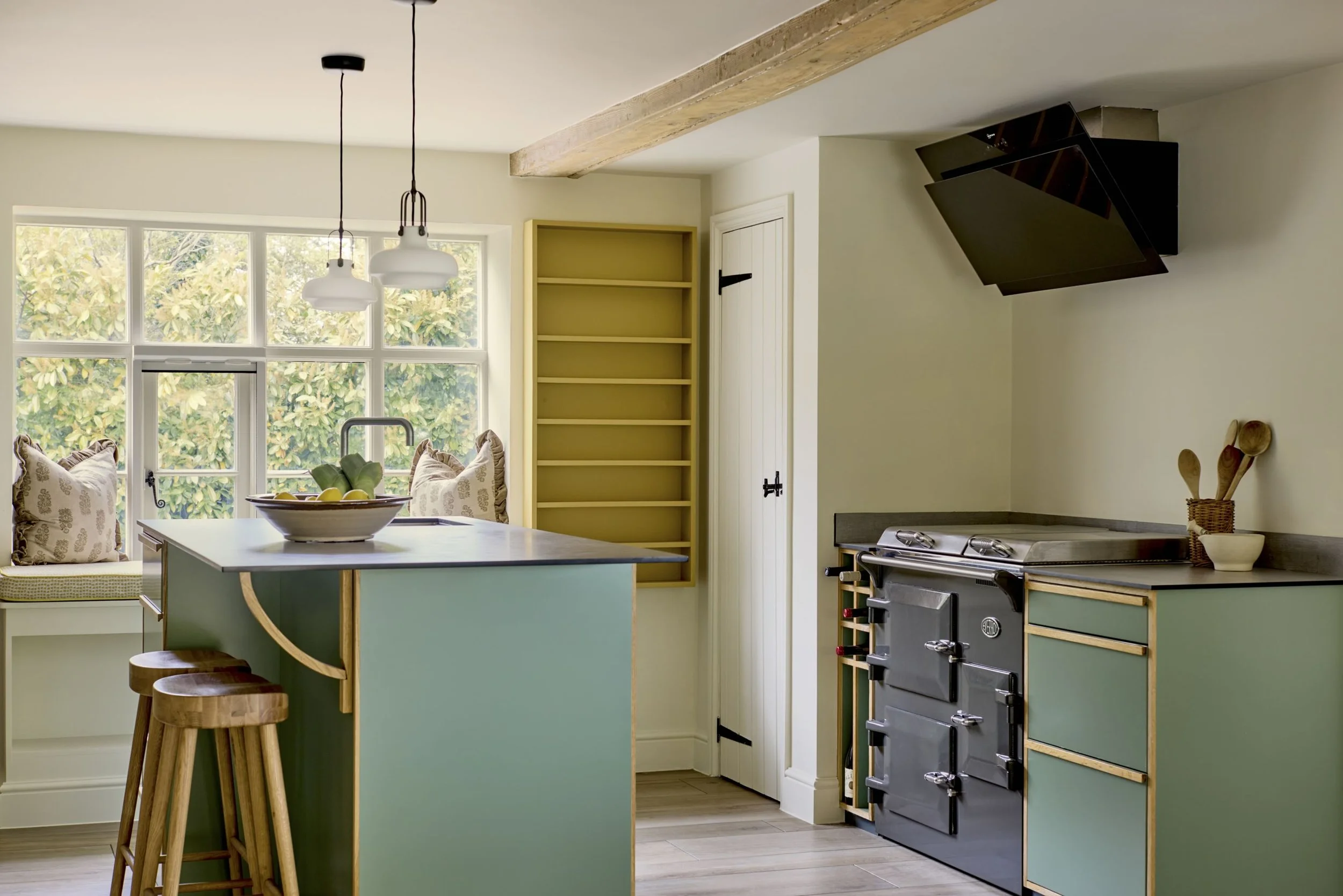 A modern kitchen with a green island and wooden stools, large windows with greenery outside, a yellow bookshelf, a black stove, and black range hood.
