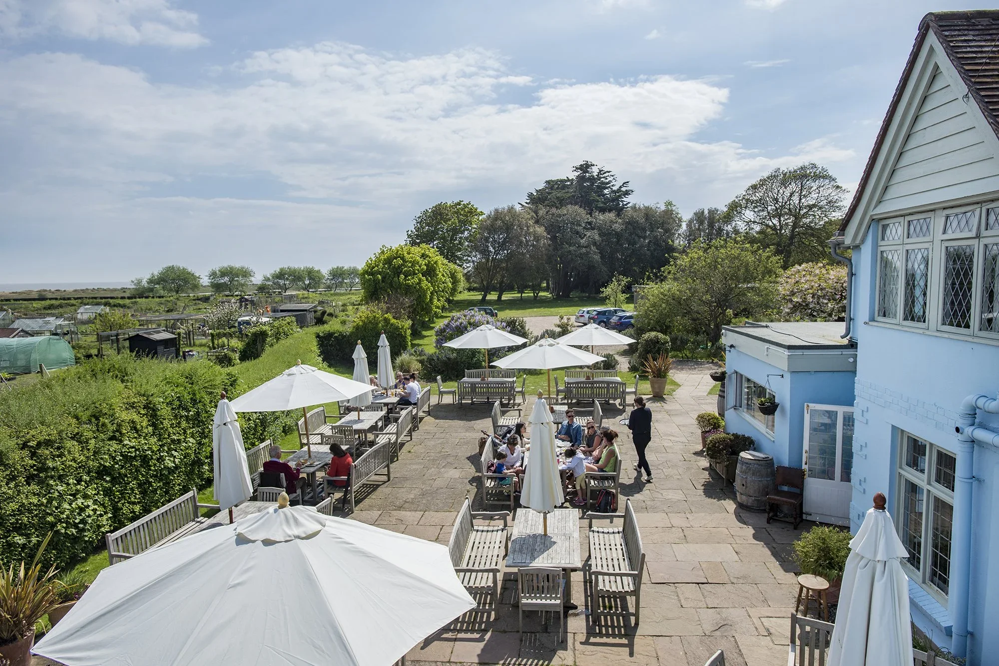 Outdoor dining area with tables, umbrellas, and people eating, adjacent to a white building with blue trim, surrounded by greenery and trees, under a partly cloudy sky.