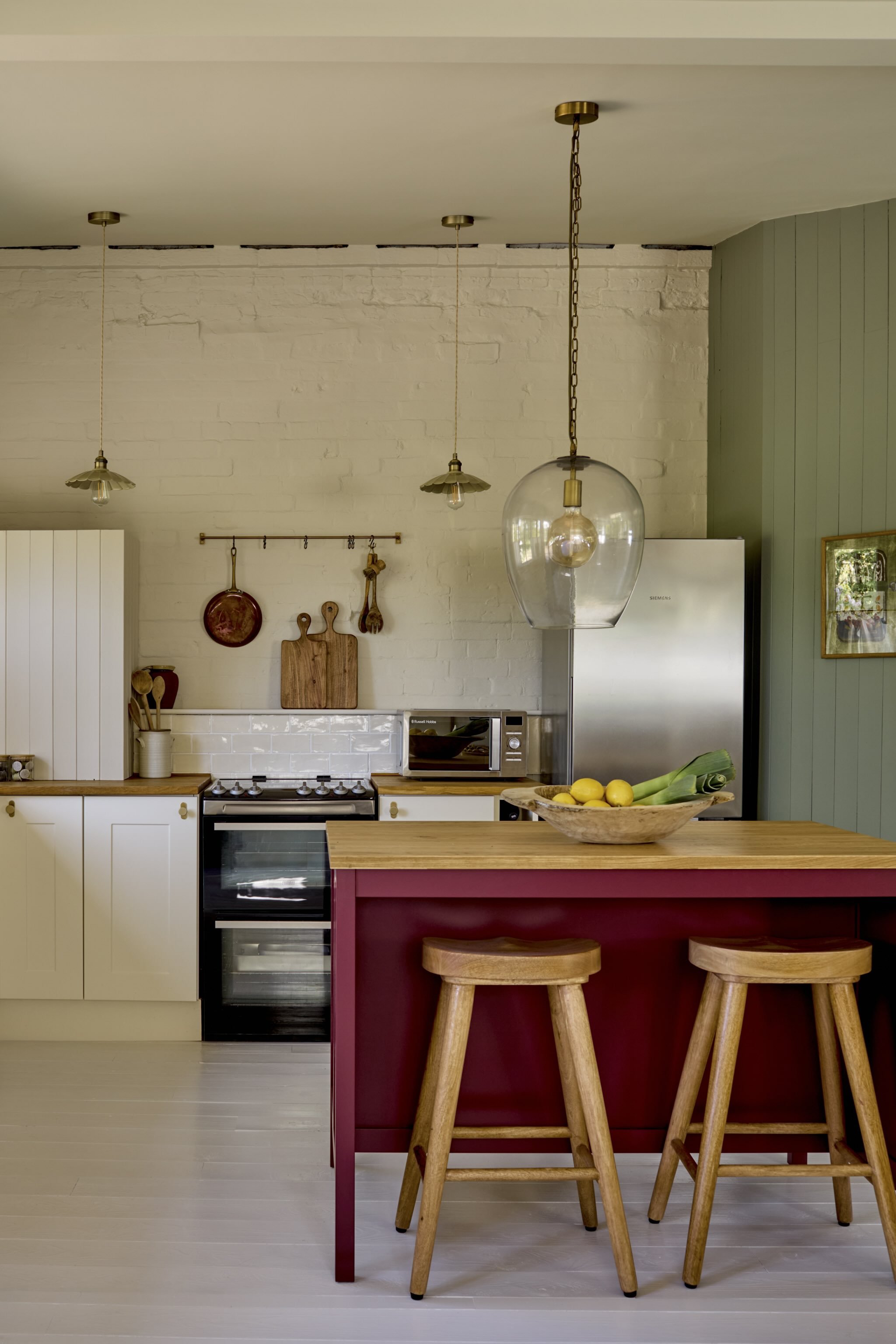 A modern kitchen with a white brick wall, green paneling, and a white ceiling. There are three hanging pendant lights and a large glass pendant lamp above a kitchen island with a wooden top. The island has a red base and three wooden stools. On the i