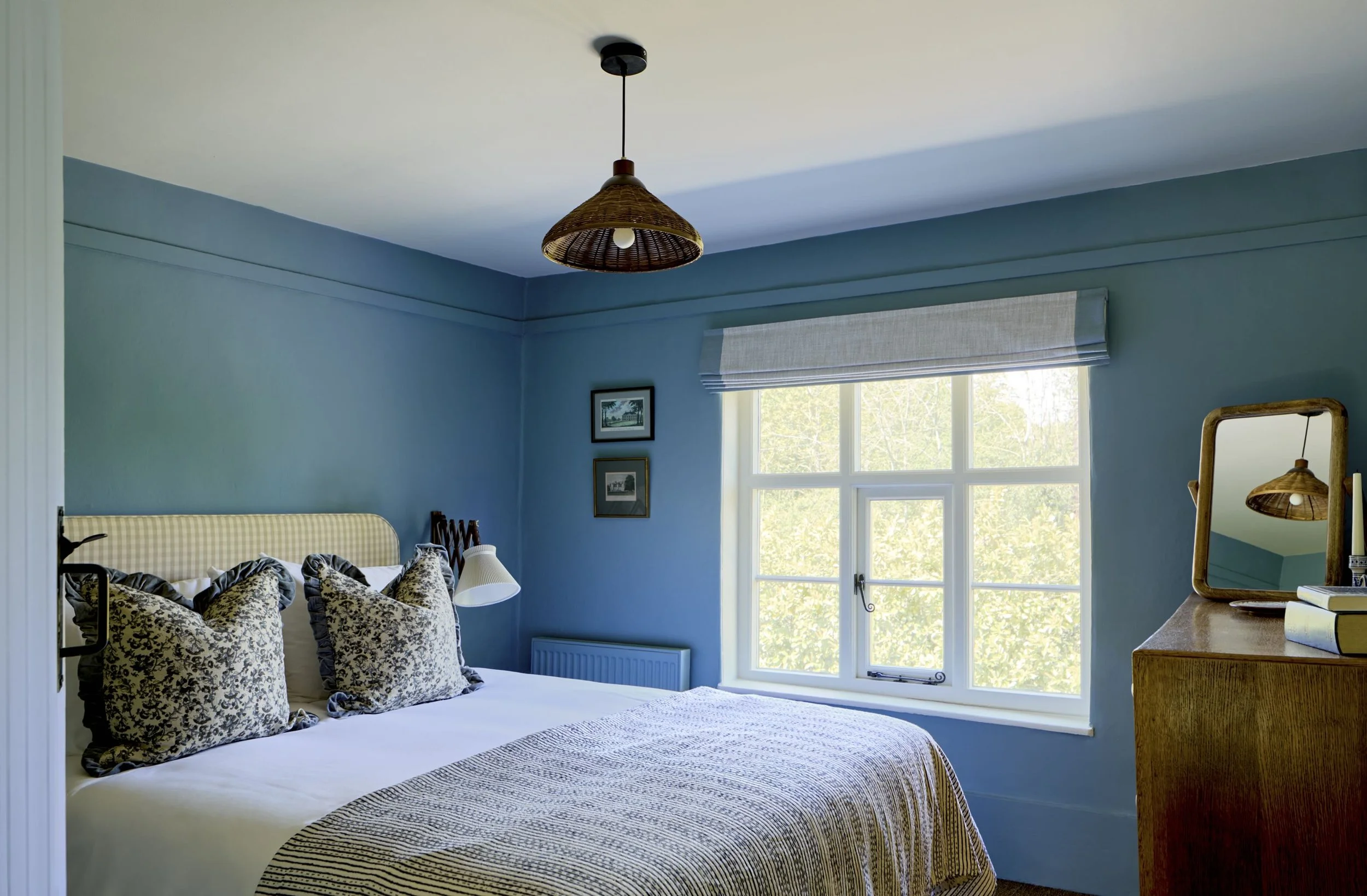 A bedroom with blue walls, a large window with a light-colored shade, a bed with floral-patterned pillows, a small white lamp, and a wicker ceiling light fixture.