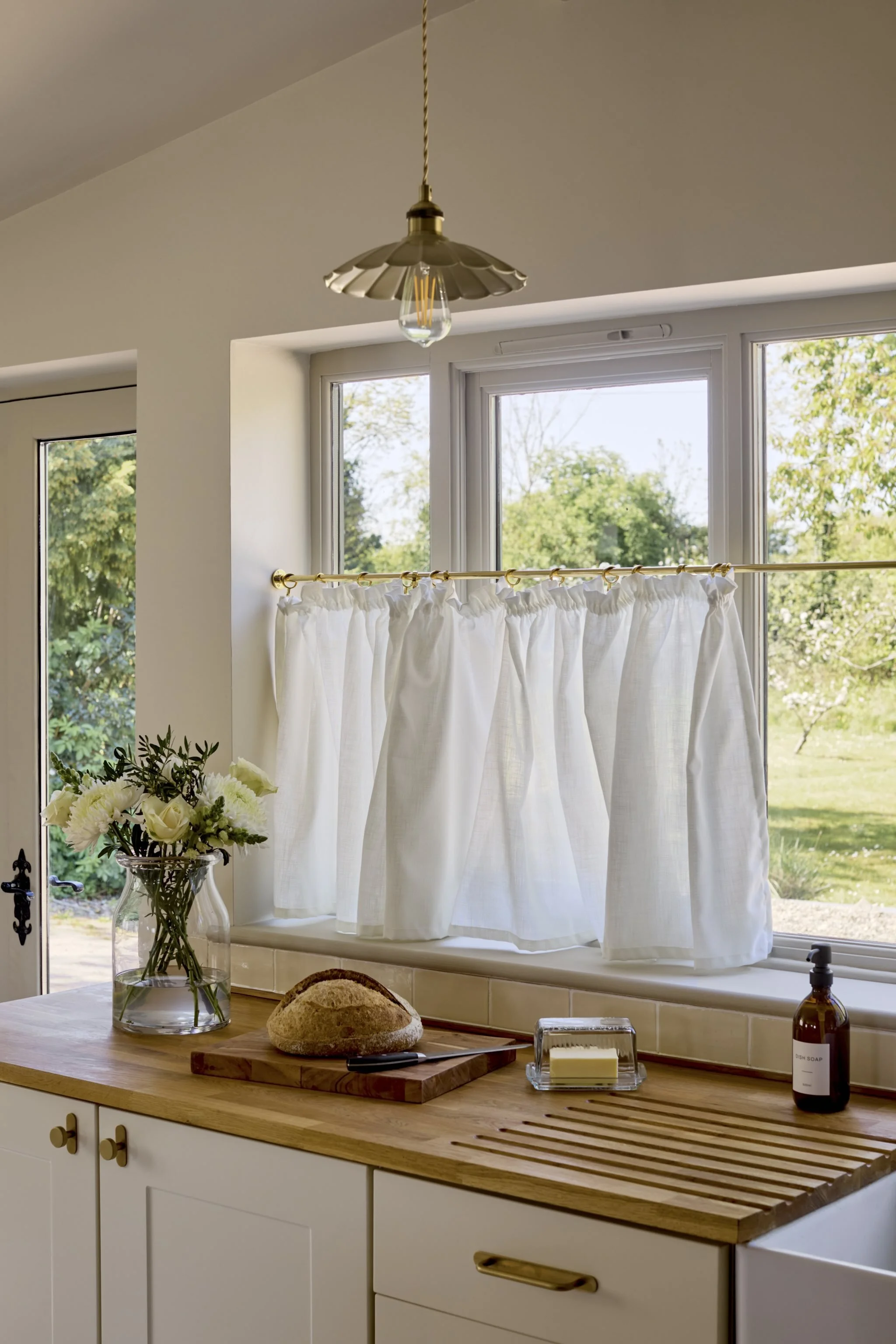 A kitchen window with white curtains, a vase with white flowers on a wooden counter, a loaf of bread on a cutting board, and soap and hand sanitizer bottles.
