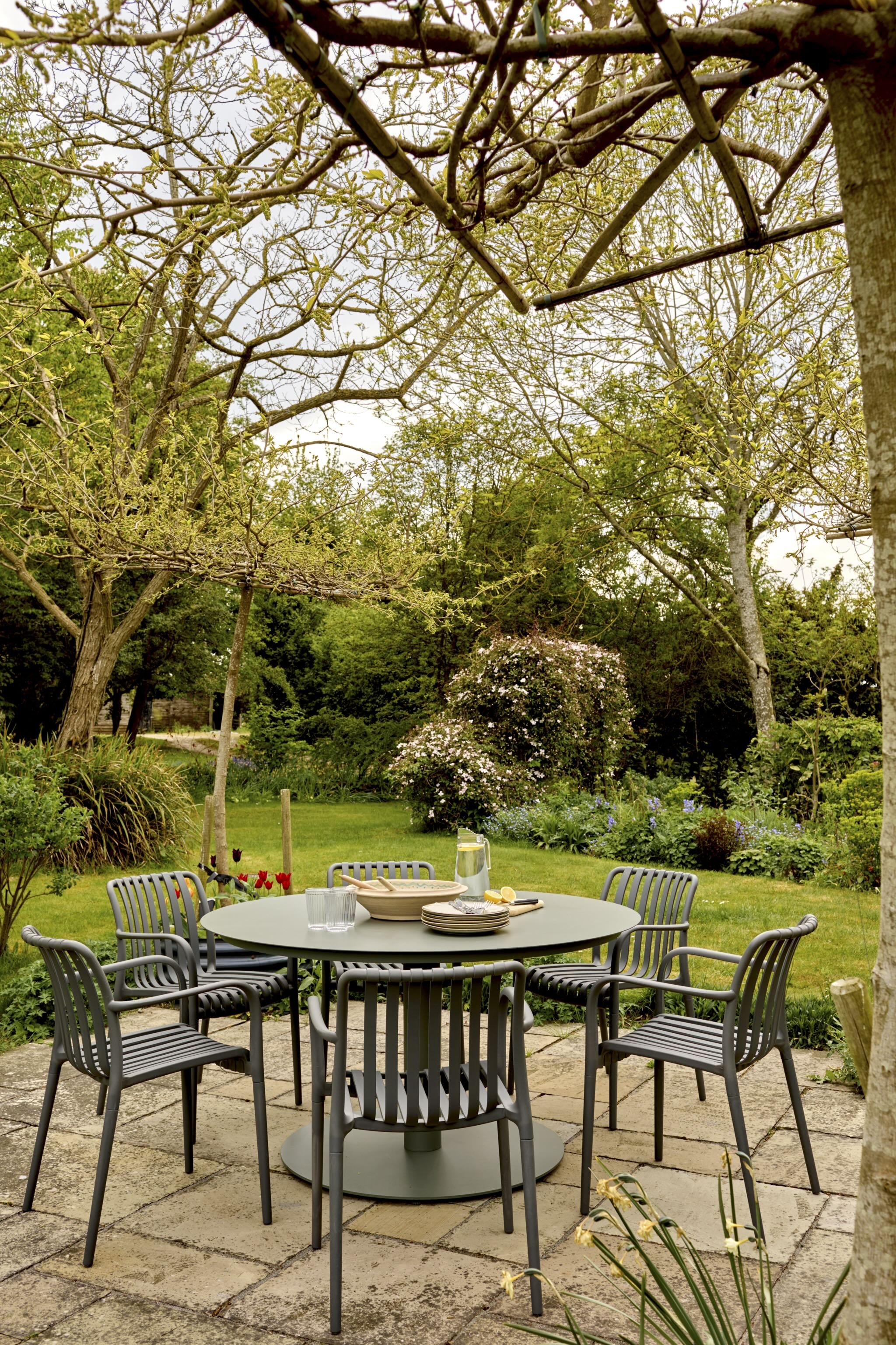 Outdoor dining area with a round table set with dishes, glasses, and a pitcher, surrounded by eight chairs, in a garden with trees, bushes, and flowers.