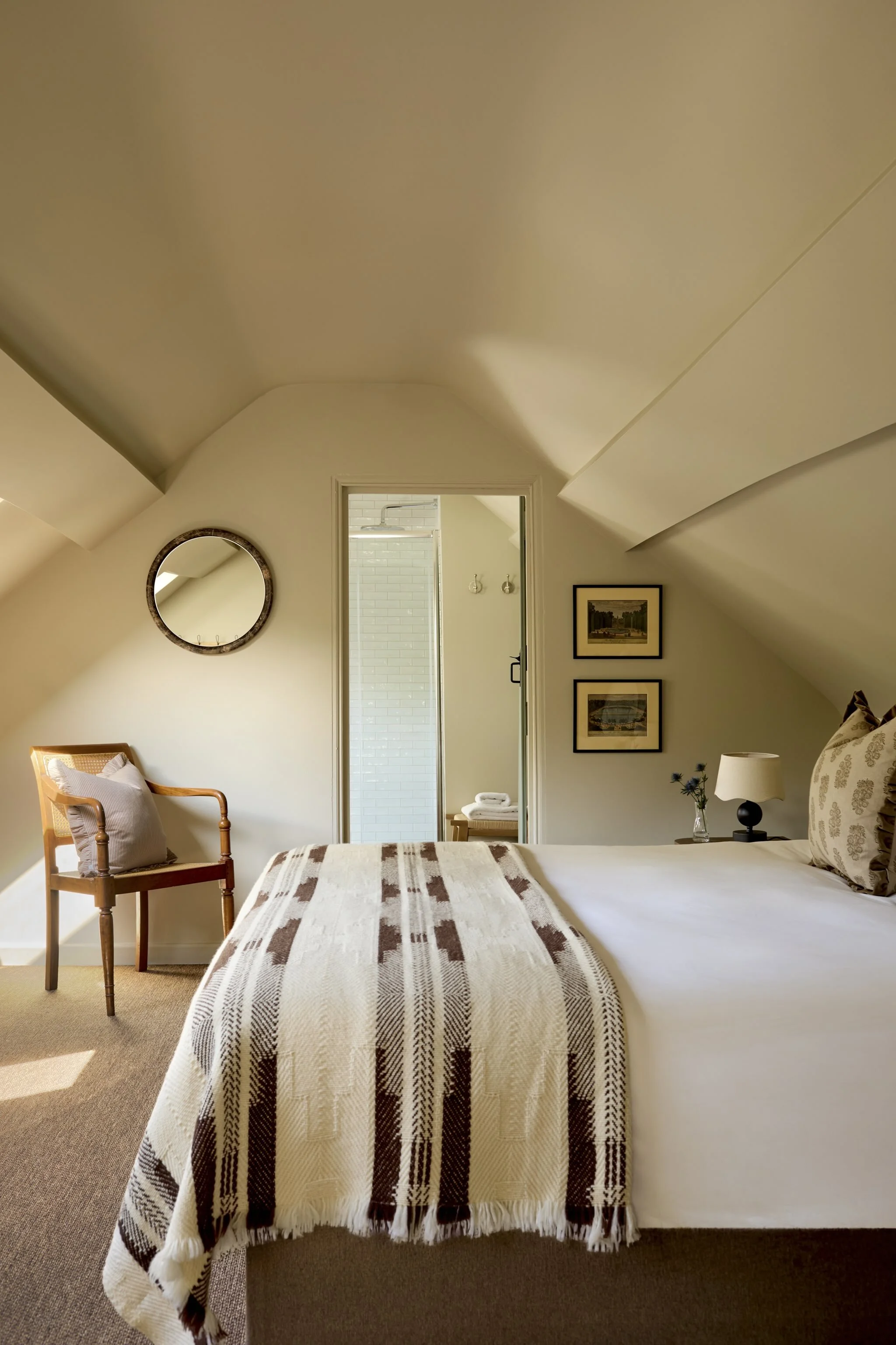 A cozy bedroom with a bed covered with a beige and brown plaid blanket, a wooden chair with a pillow, wall art, a mirror, and a nightstand with a lamp, chair, and framed pictures, with an open door leading to a walk-in shower in the background.