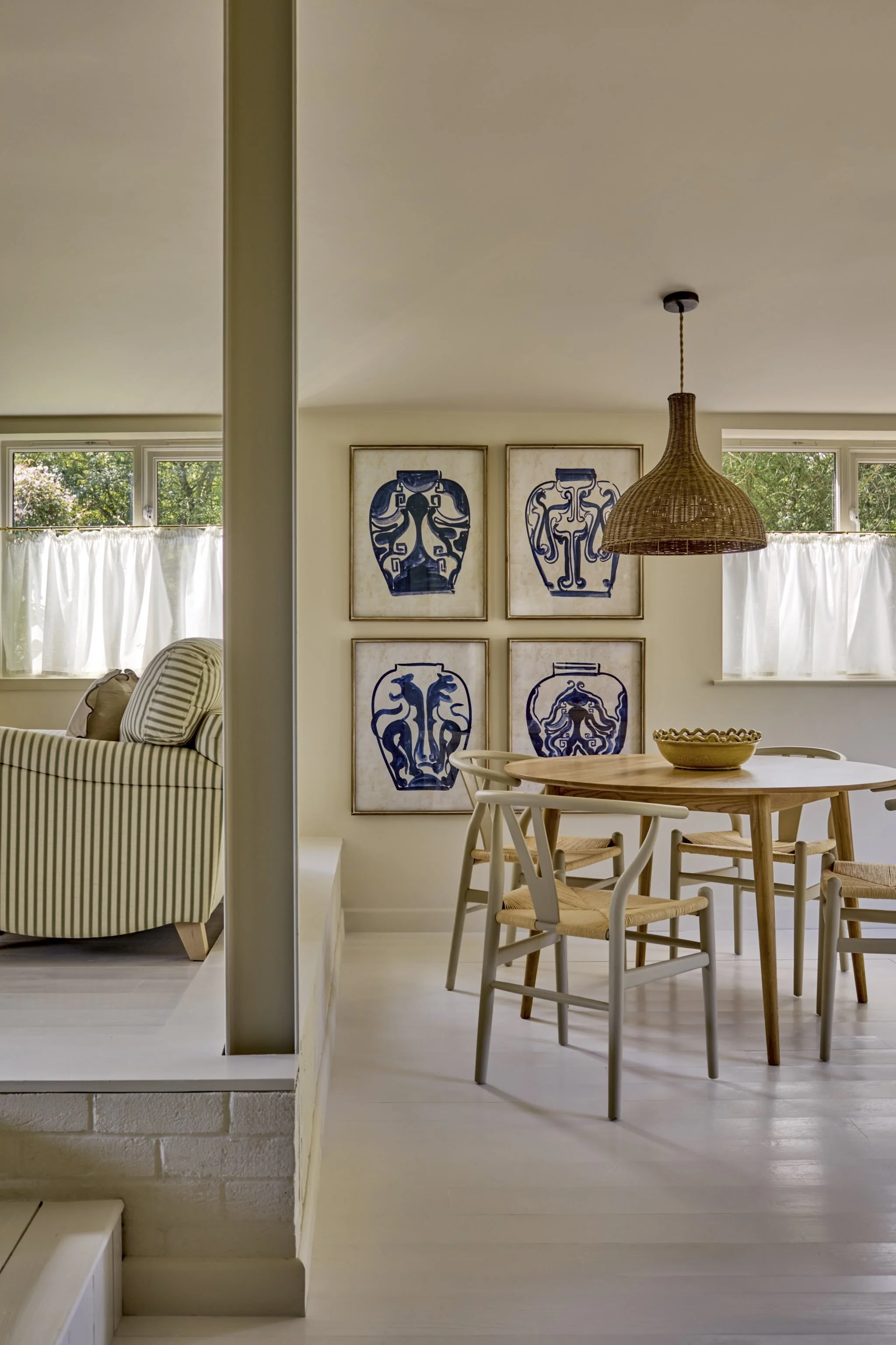 A view of a room with a dining table and four chairs, a wall with four framed art pieces featuring blue pottery, a woven pendant light, and windows with white curtains, natural light illuminating the space.