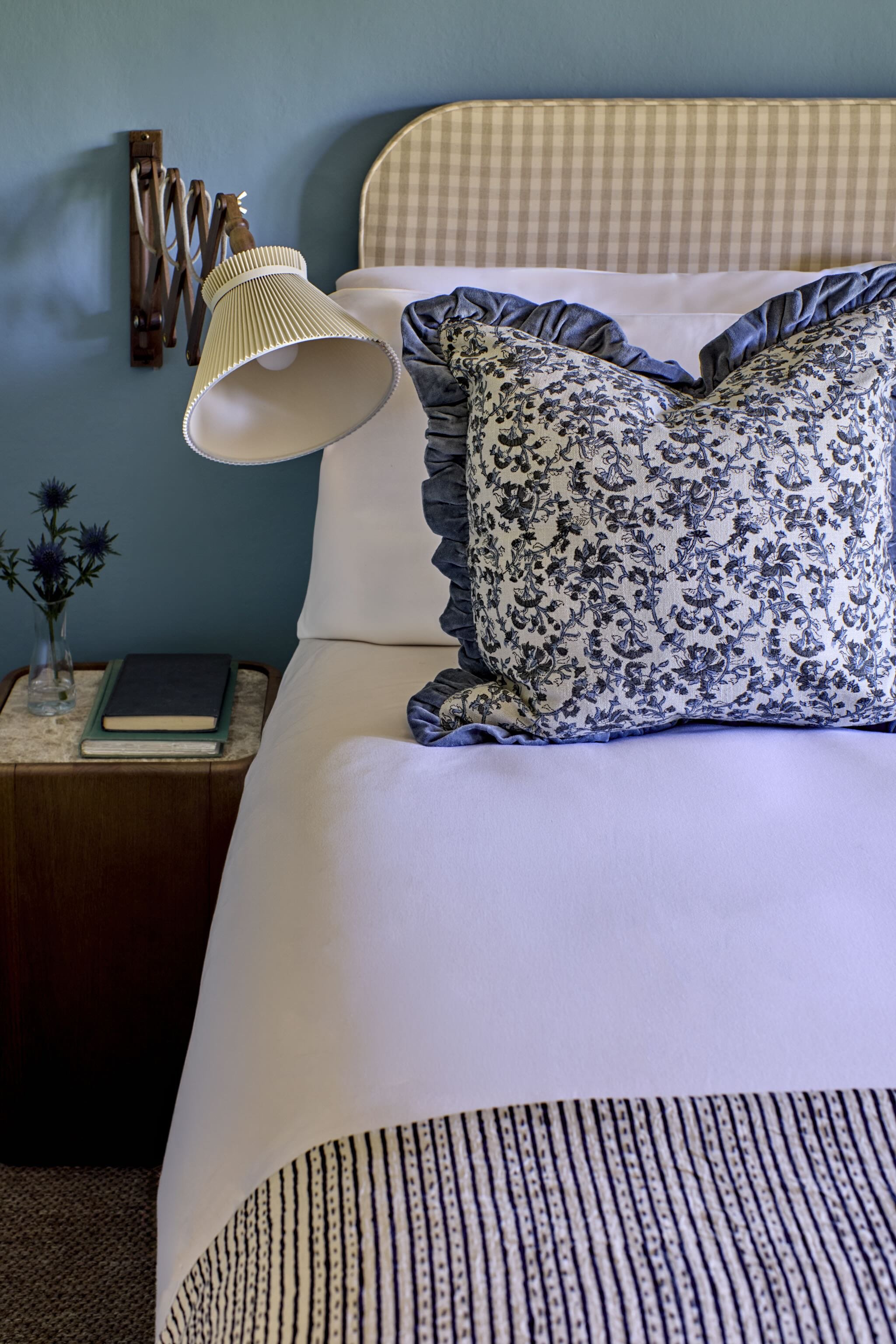 Close-up of a bed with pillows and bedspread, next to a nightstand with books, a glass vase with purple flowers, and a booked, with a lamp and a blue wall in the background.