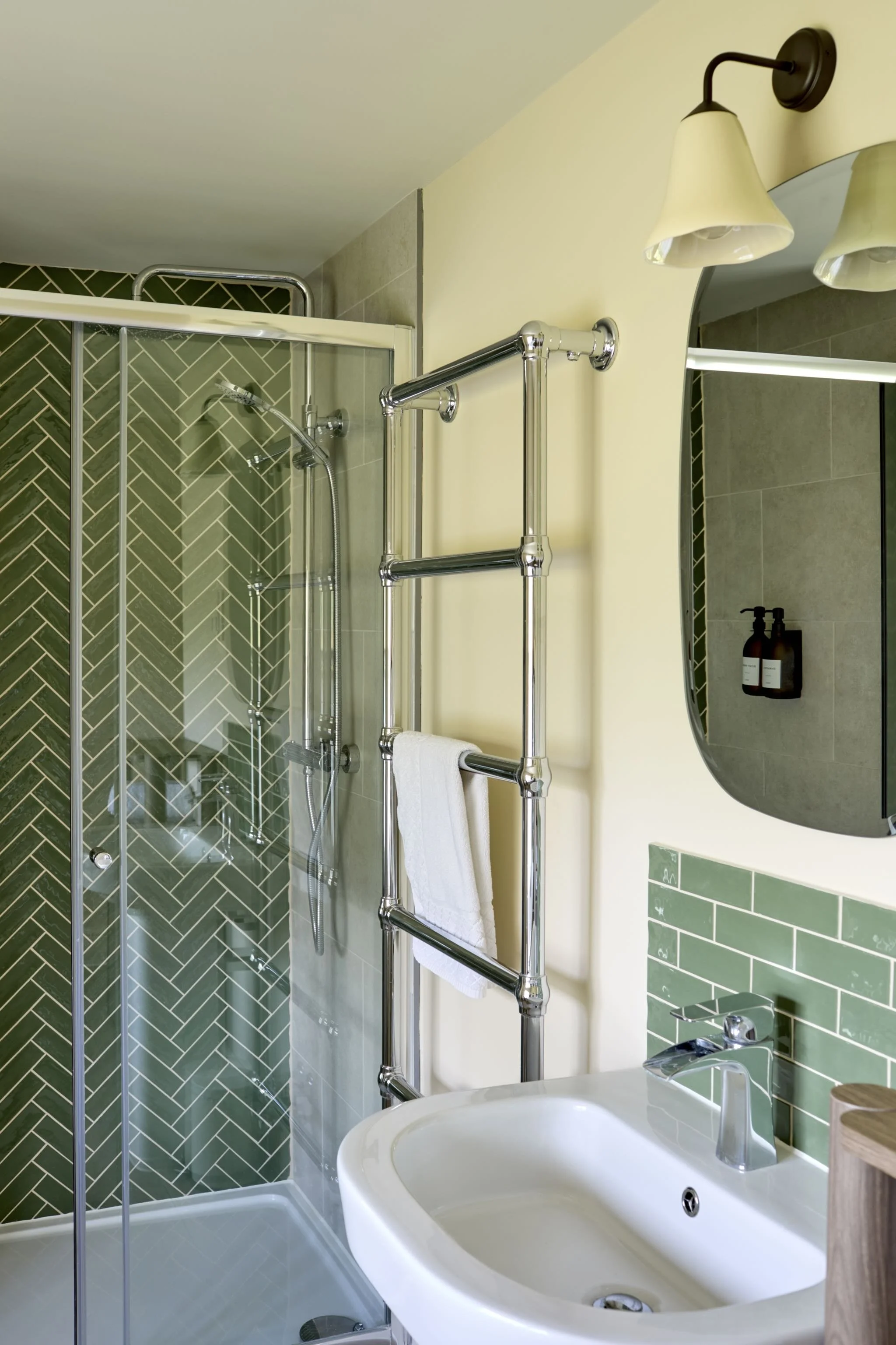 A bathroom with a walk-in shower with glass doors, green herringbone tile, a chrome towel warmer with a white towel, a white sink with a modern faucet, a mirror, beige walls, and two wall-mounted lamps with cream-colored shades. Two bottles of liquid