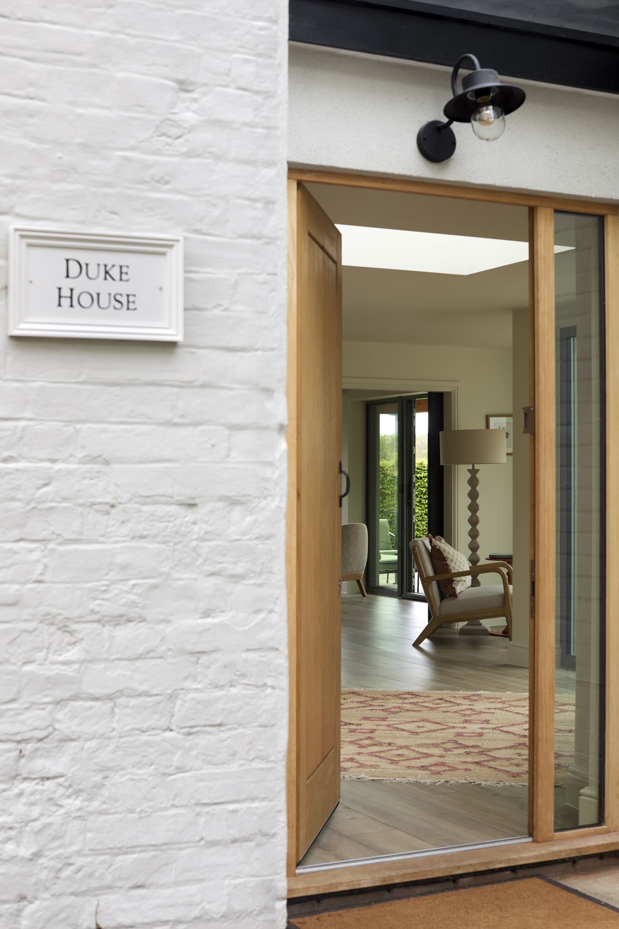 The image shows the entrance to a house with a white brick wall and a wooden-framed sliding glass door. Inside, there are chairs, a lamp, and a view of a backyard with green trees.