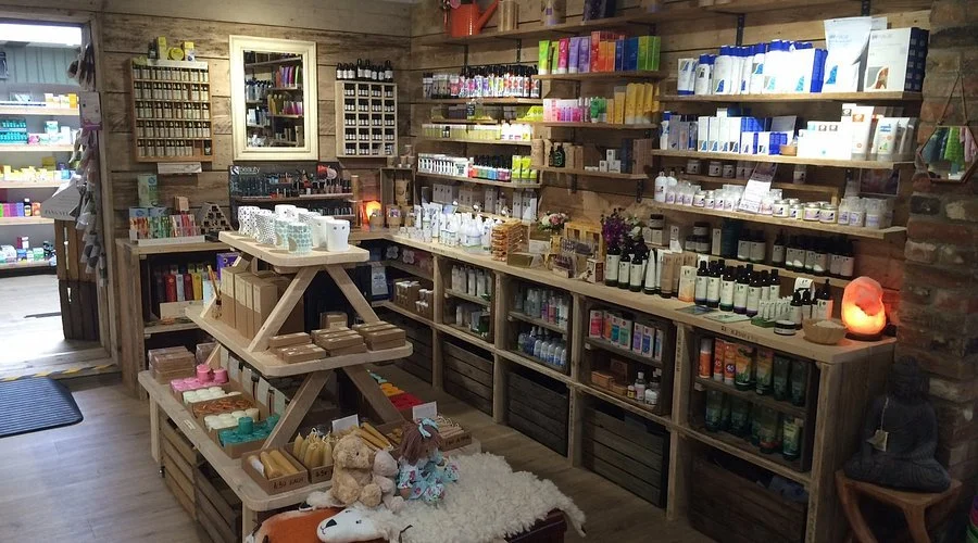 Inside a natural health and beauty store with wooden shelves stocked with skincare, haircare, and wellness products. There are shelves on the walls, a display table in the center with various items, and cozy decor including salt lamps and a small Buddha statue.